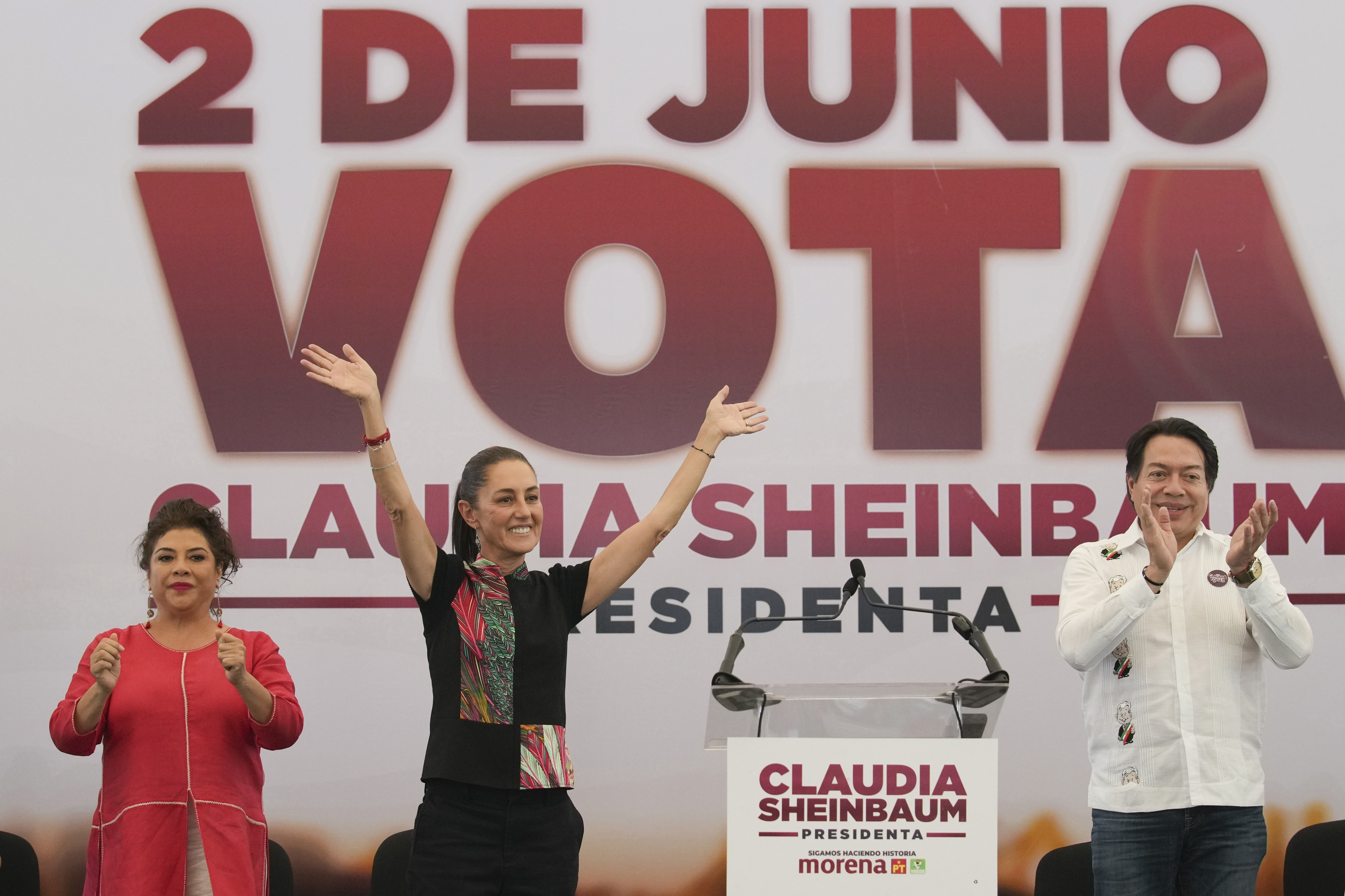 Claudia Sheinbaum raises her arms in front of a sign that says "2 de junio vota"