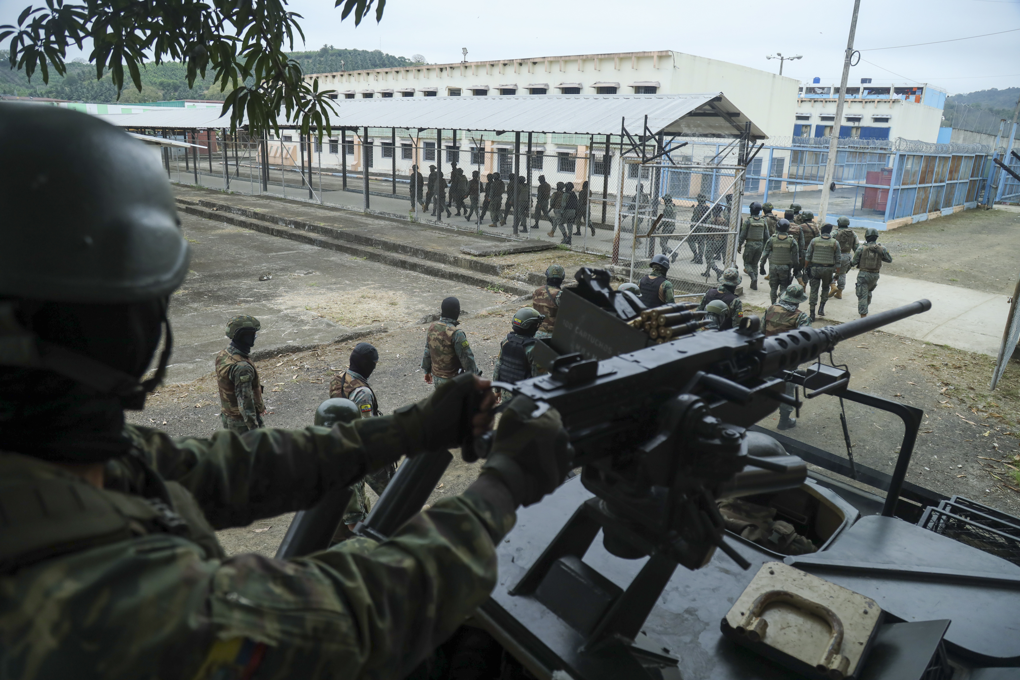 A soldier sits behind the sight of a rifle, looking out on the El Rodeo prison, its long white buildings and chain-link fence in the distance.