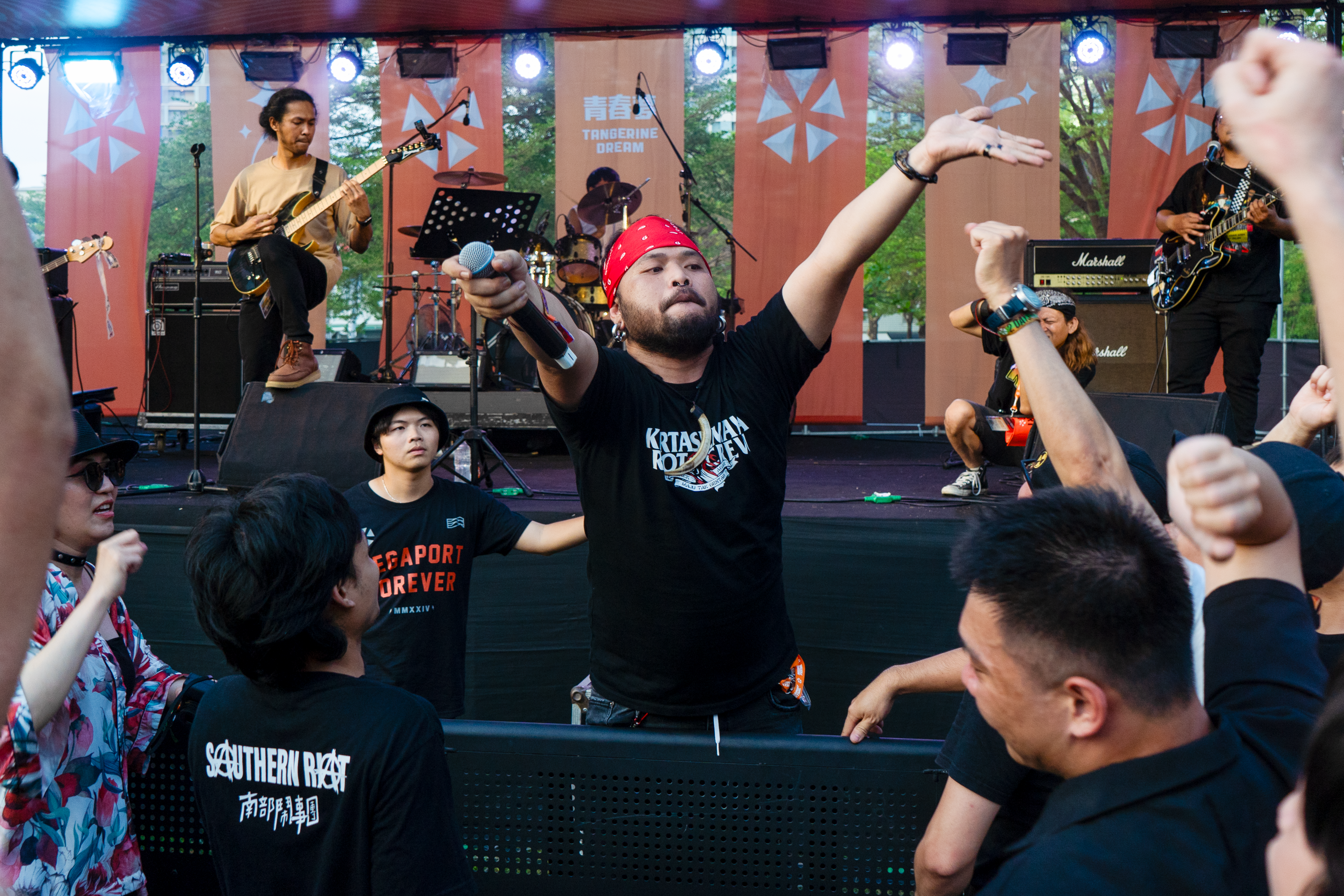 Southern Riot's lead singer Rudi on stage. He's wearing a black T-shirt and a red bandana around his head. The other band members are behind. There are crowds around him. He is holding the microphone out to the fans