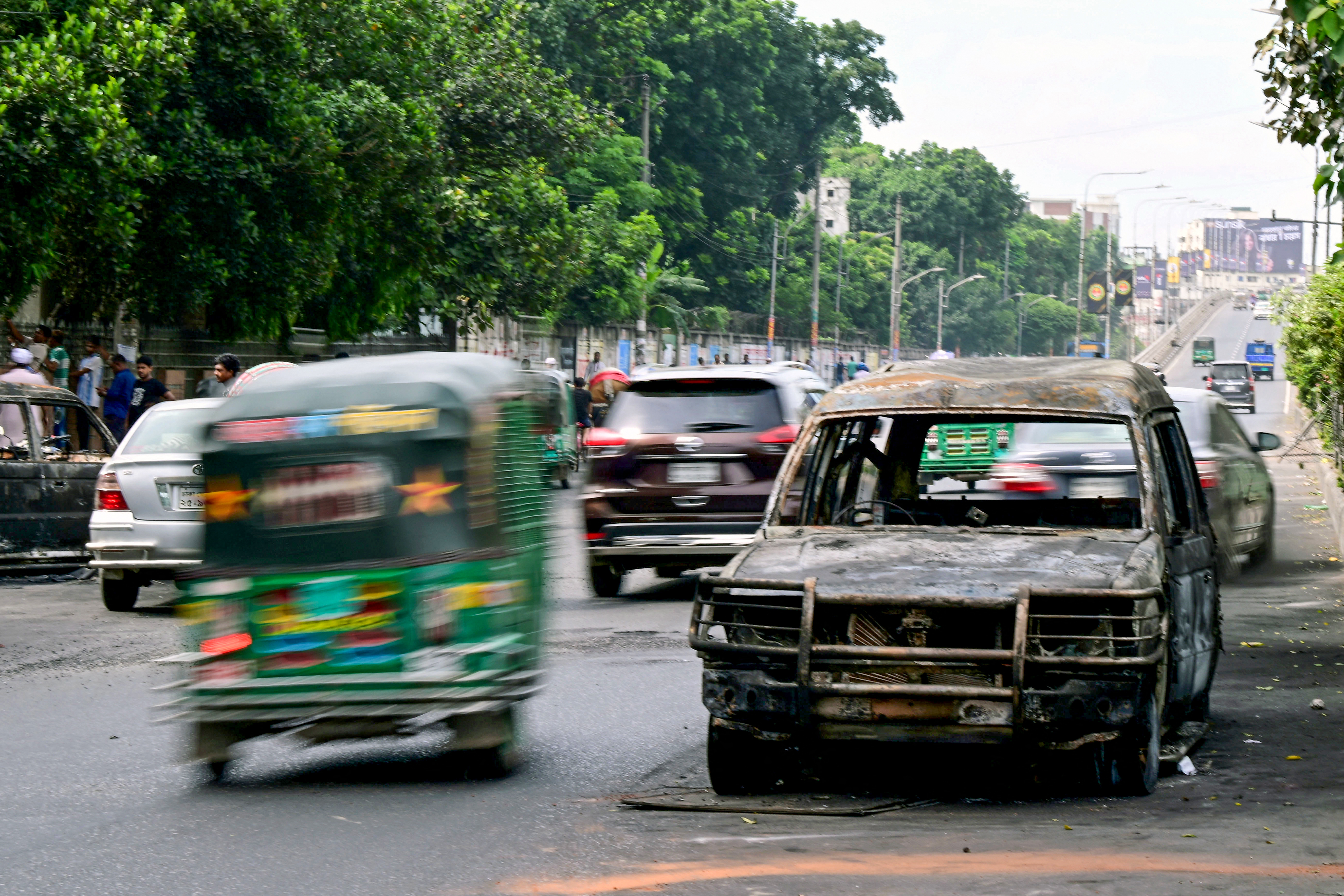 Commuters drive past a burnt car after students set it on fire amid the ongoing anti-quota protest in Dhaka