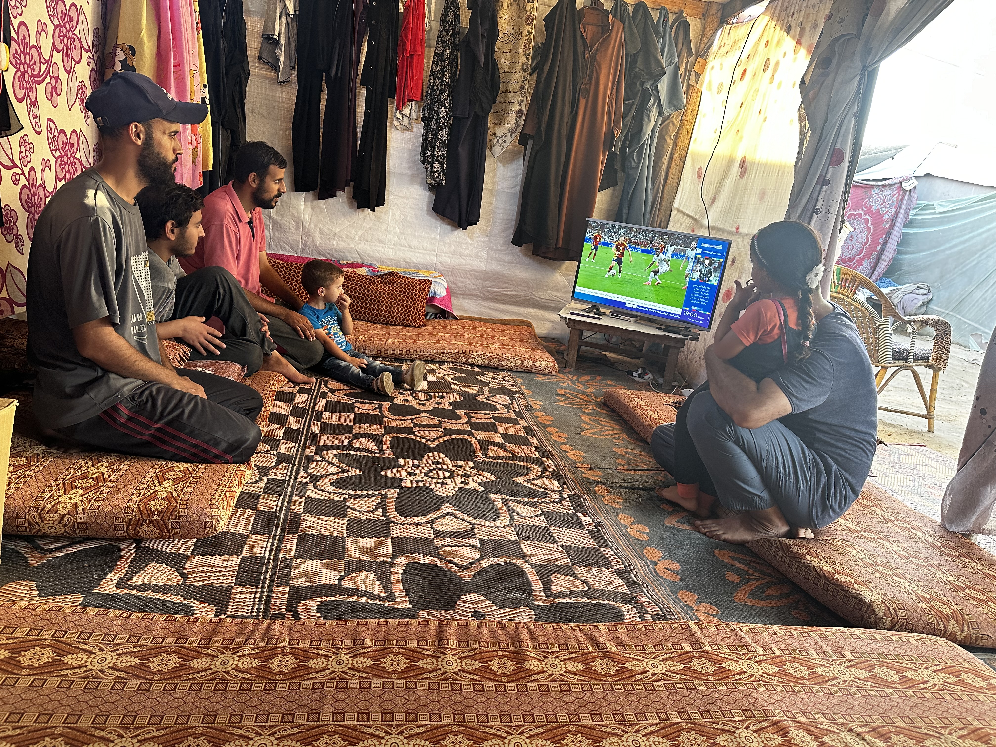 Gaza residents Omar Ghayyad (top left), with his son, and Mohammed Ghayyad, with his daughter (right), watch a Euro 2024 football match in their tent in Deir el-Balah, Gaza [Abubaker Abed/Al Jazeera]