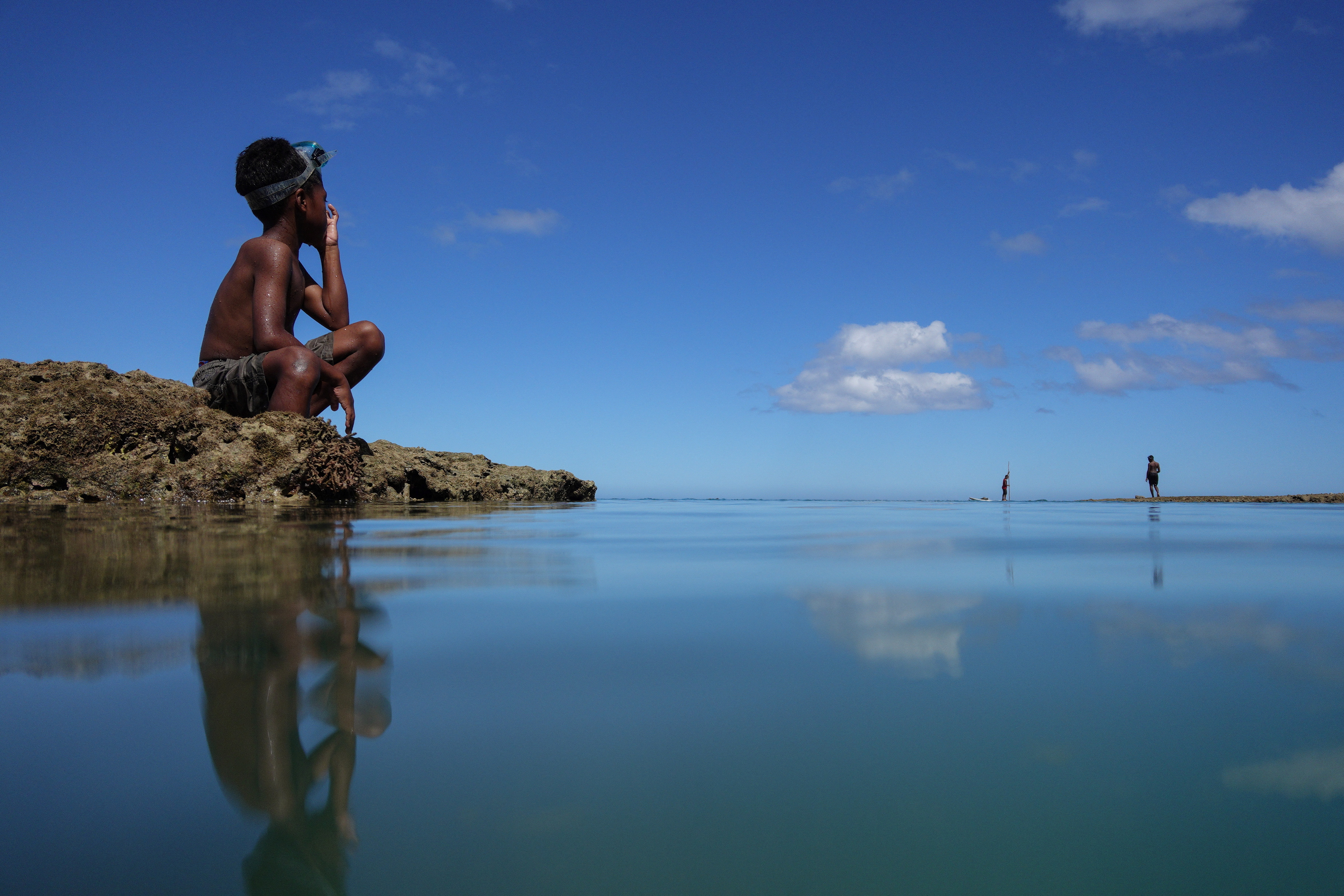 A boy squatting on rocks next to the sea in Fiji. Two other people can be seen in the distance.