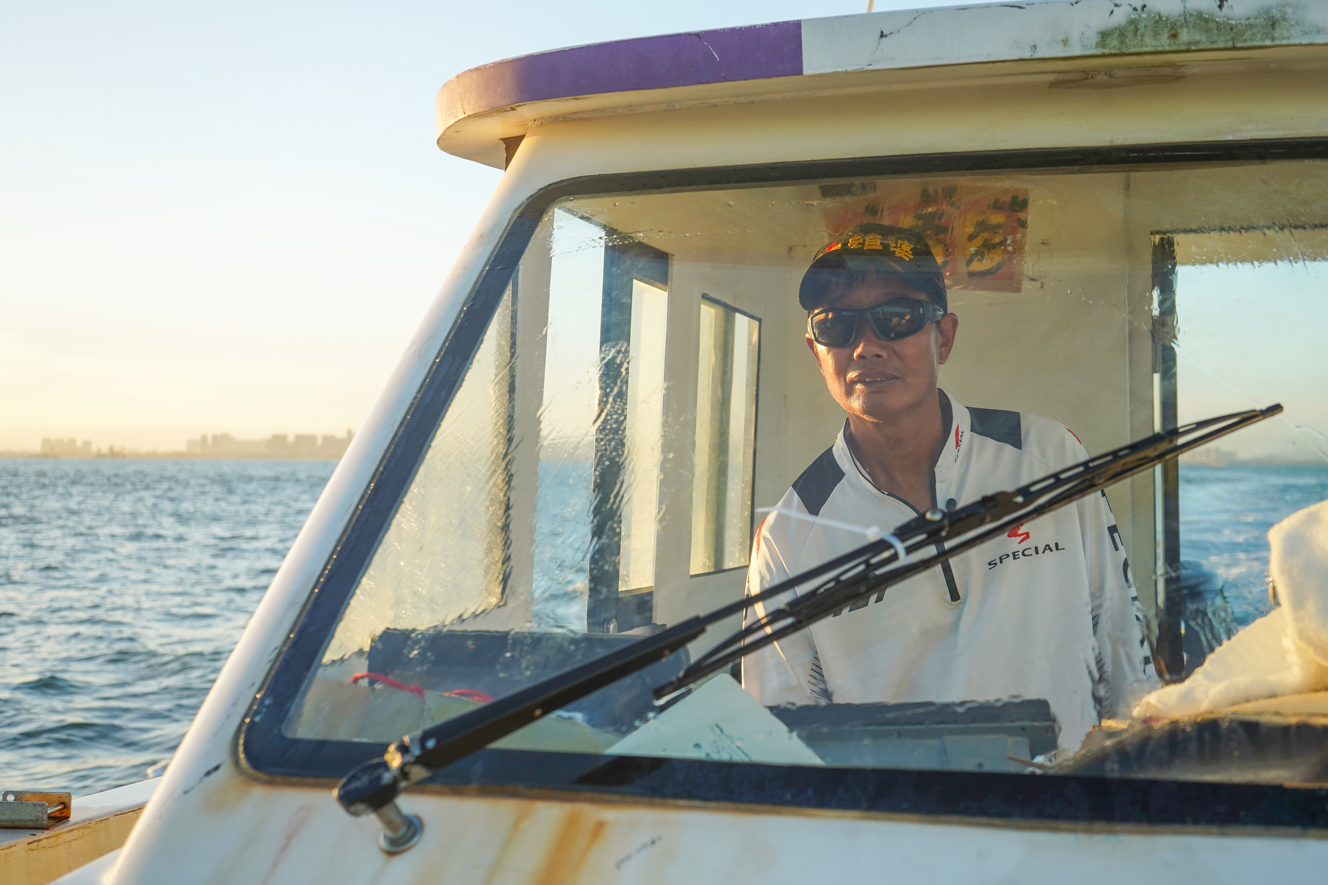 u Wen Shiung on his boat. He is standing at the wheel. He is wearing a white shirt, baseball cap and sunglasses