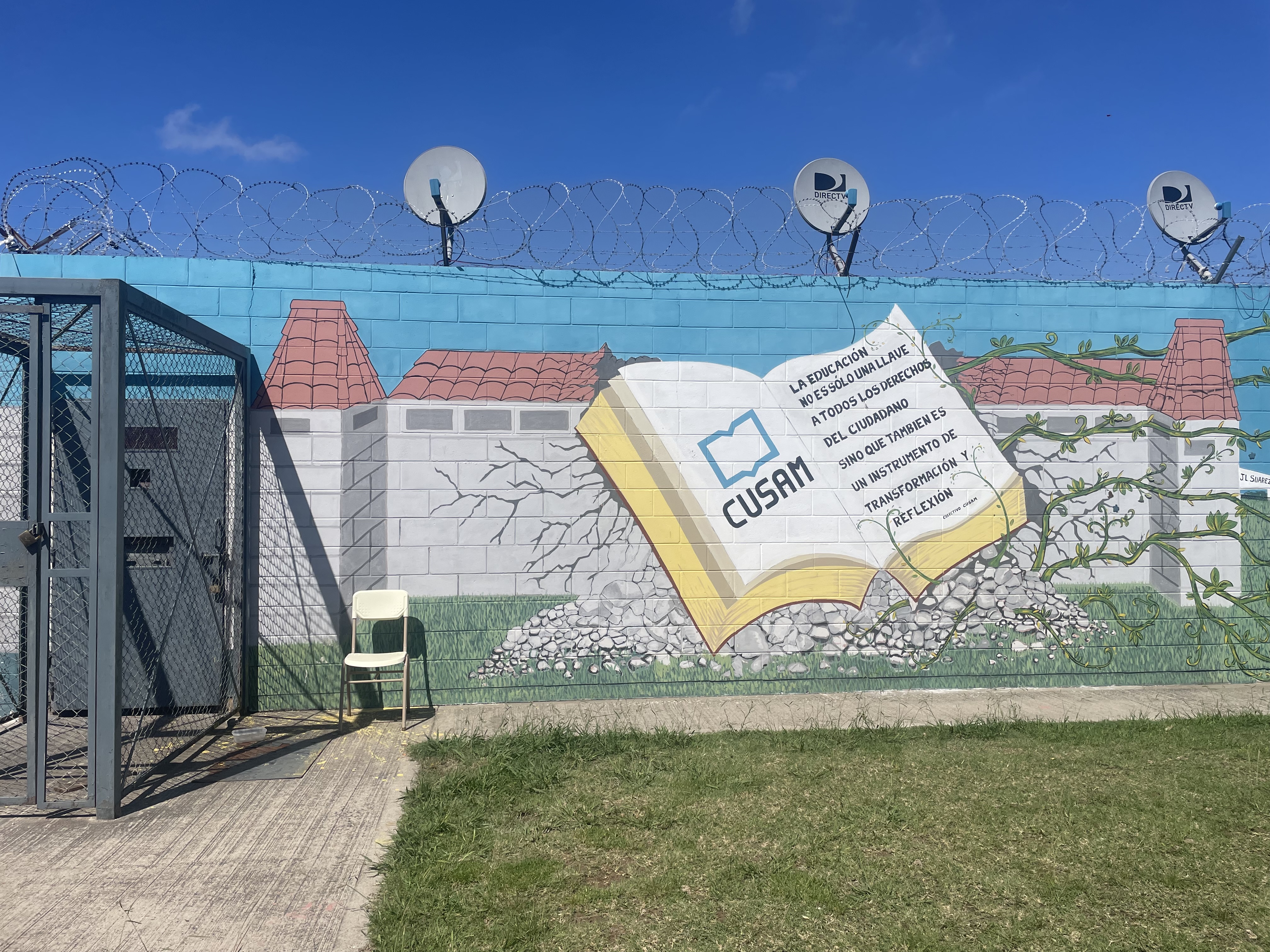 A mural on a cinderblock wall — below satellite dishes and barbed wire — shows an open book with the word "CUSAM" inside as well as a phrase: "La educacion no es solo una llave a todos los derechos del cuidadano, sino que tambien es un instrumento de transformacion y reflexion.'