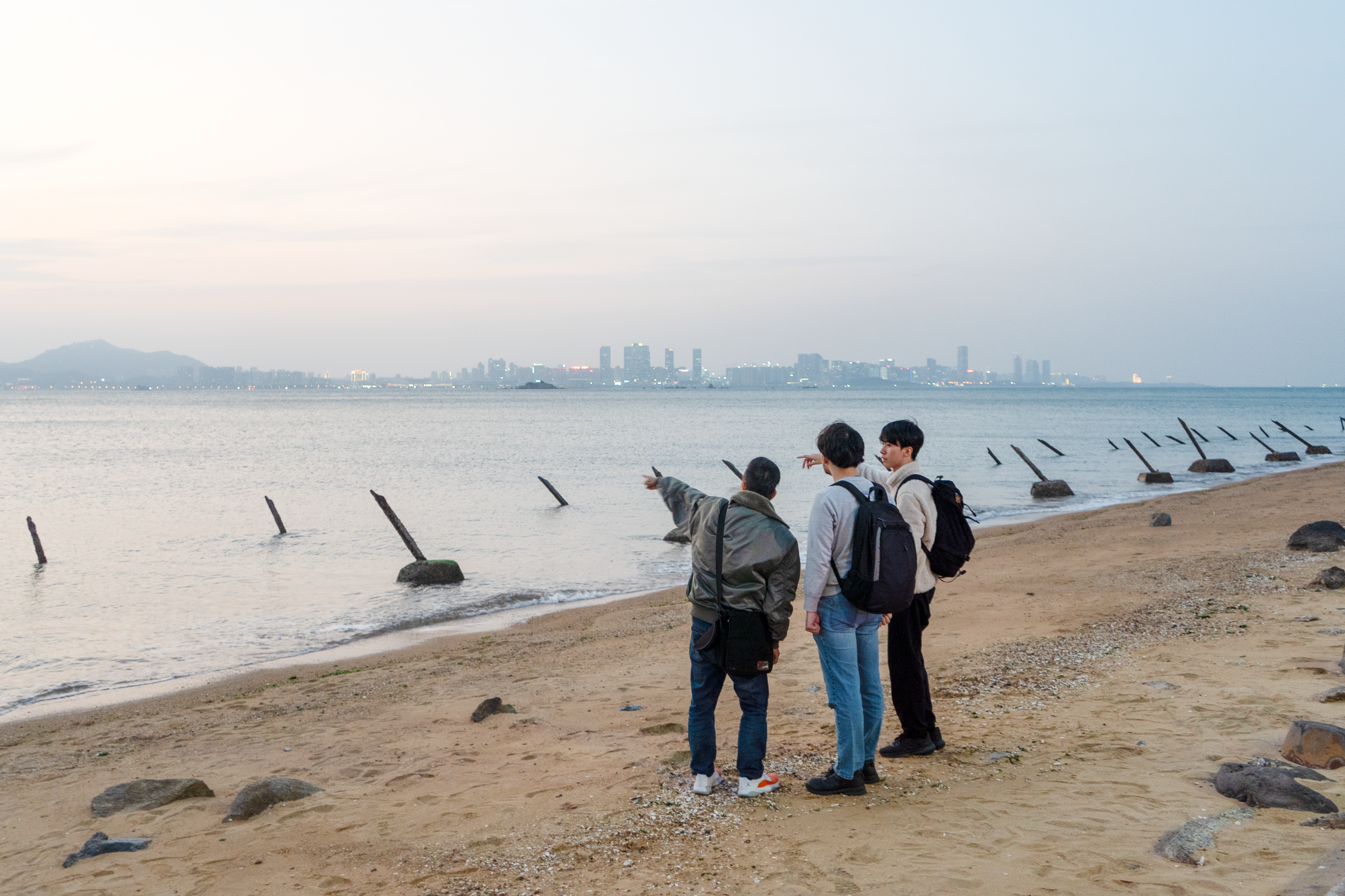 Three men on a beach in Kinmen. They are standing on the sand pointing at Xiamen which can be seen in the distance. There are anti-invasion spikes on the beach