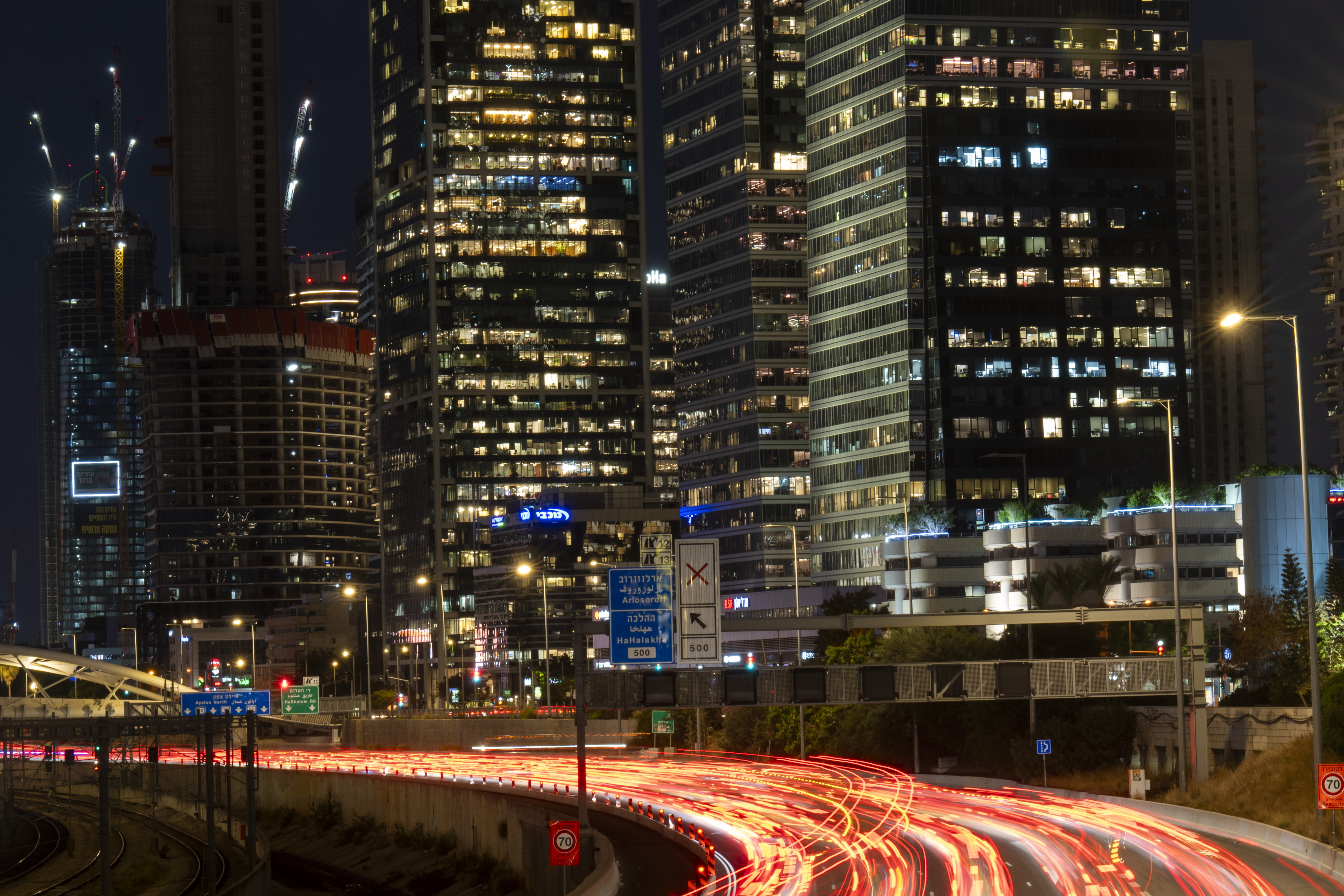 Traffic moves along a road in front of tall buildings at night