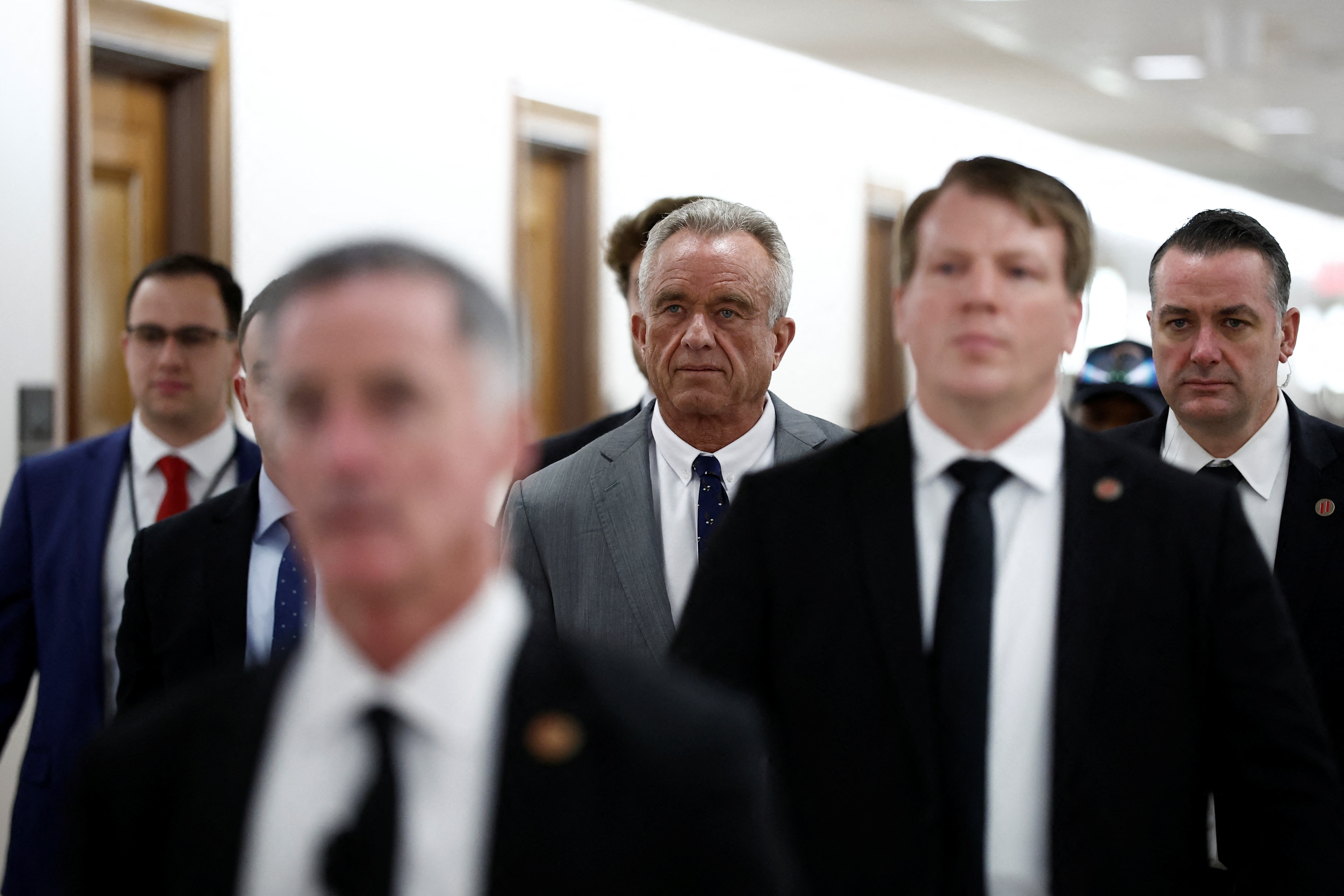 Robert F Kennedy Jr walks through the Dirksen Senate Office Building with other officials in suits and ties.