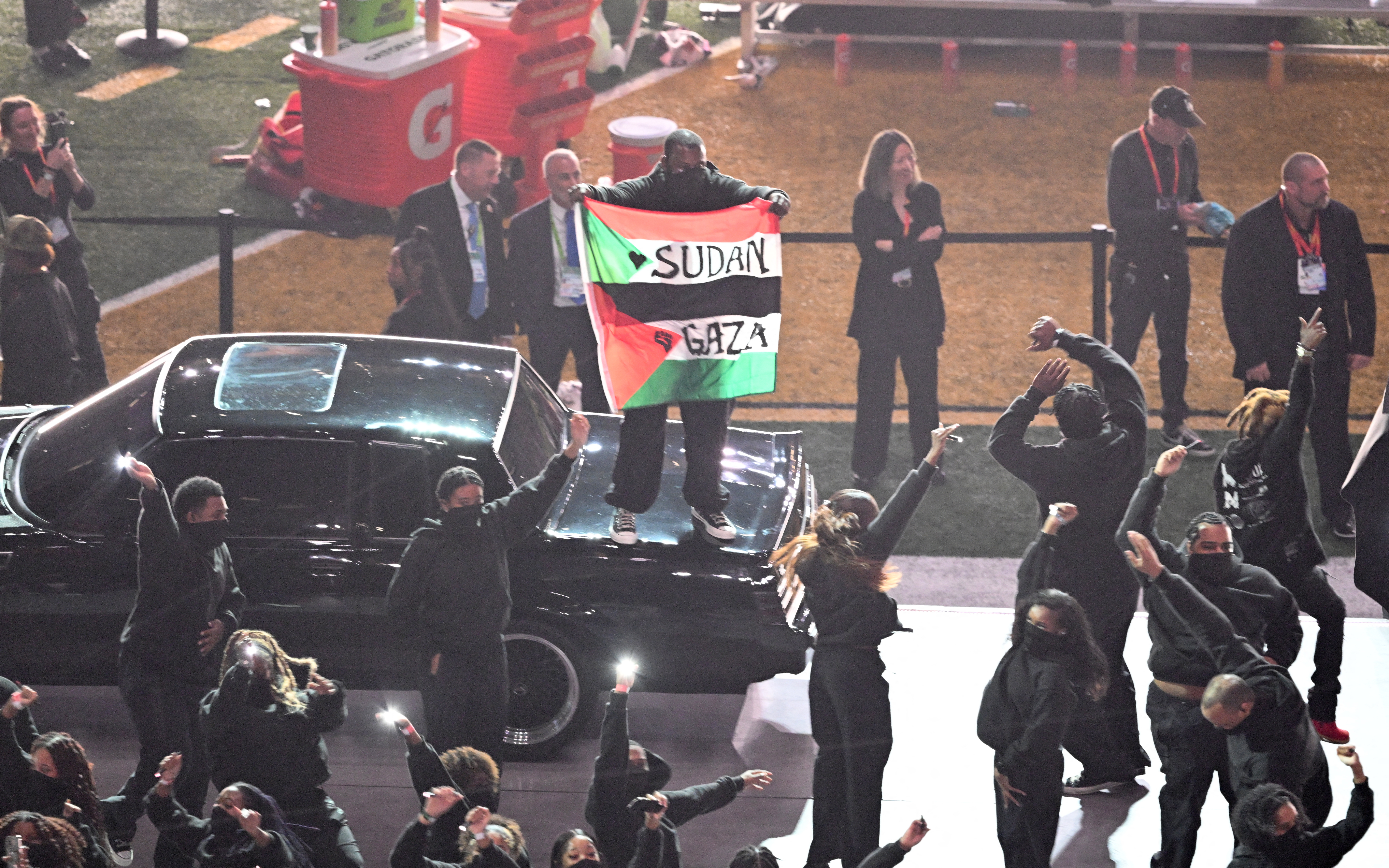 a person on the back of a black car holds.a red green and black flag that says Sudan Gaza