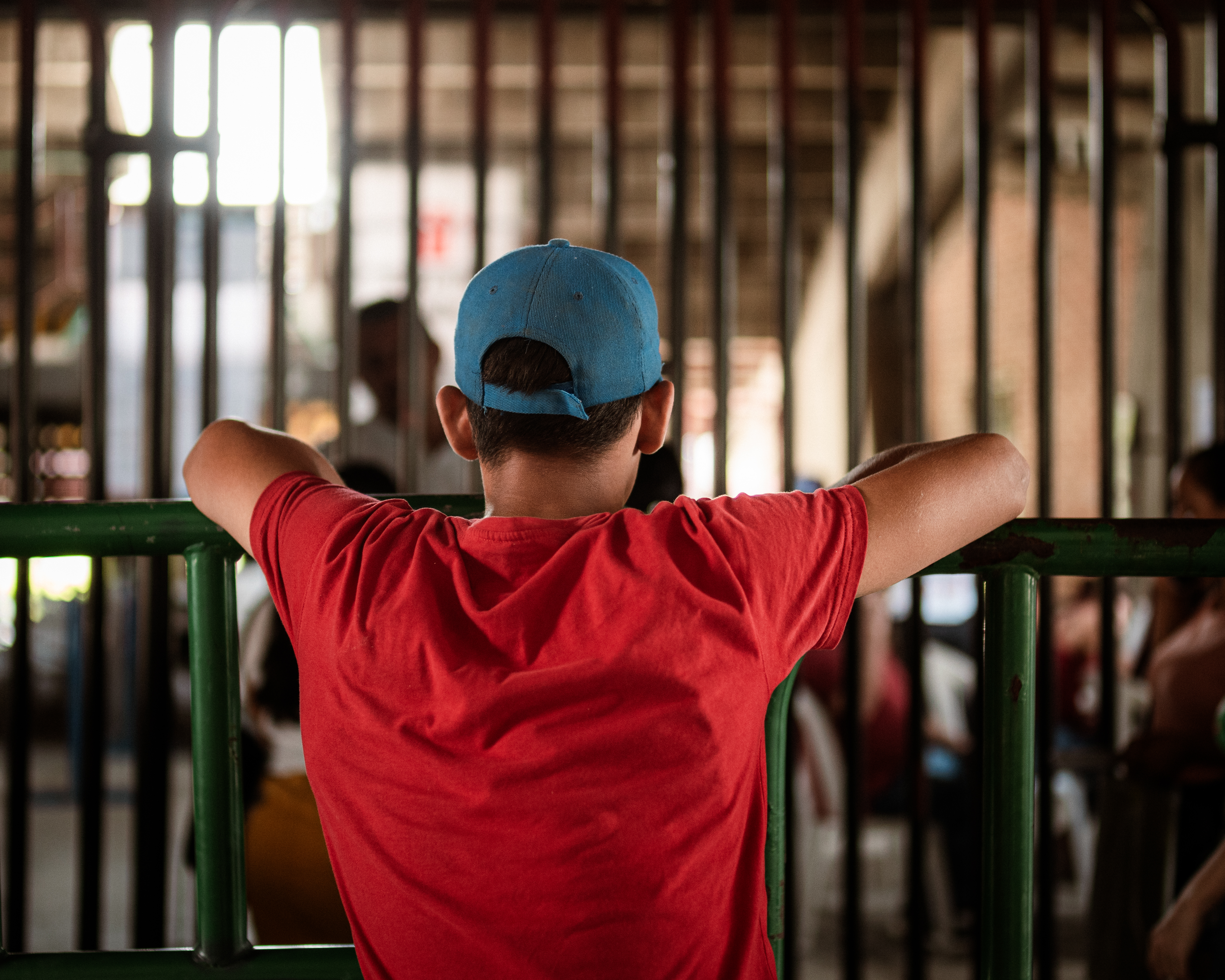 A young man in a baseball cap and a red shirt leans against a metal barricade