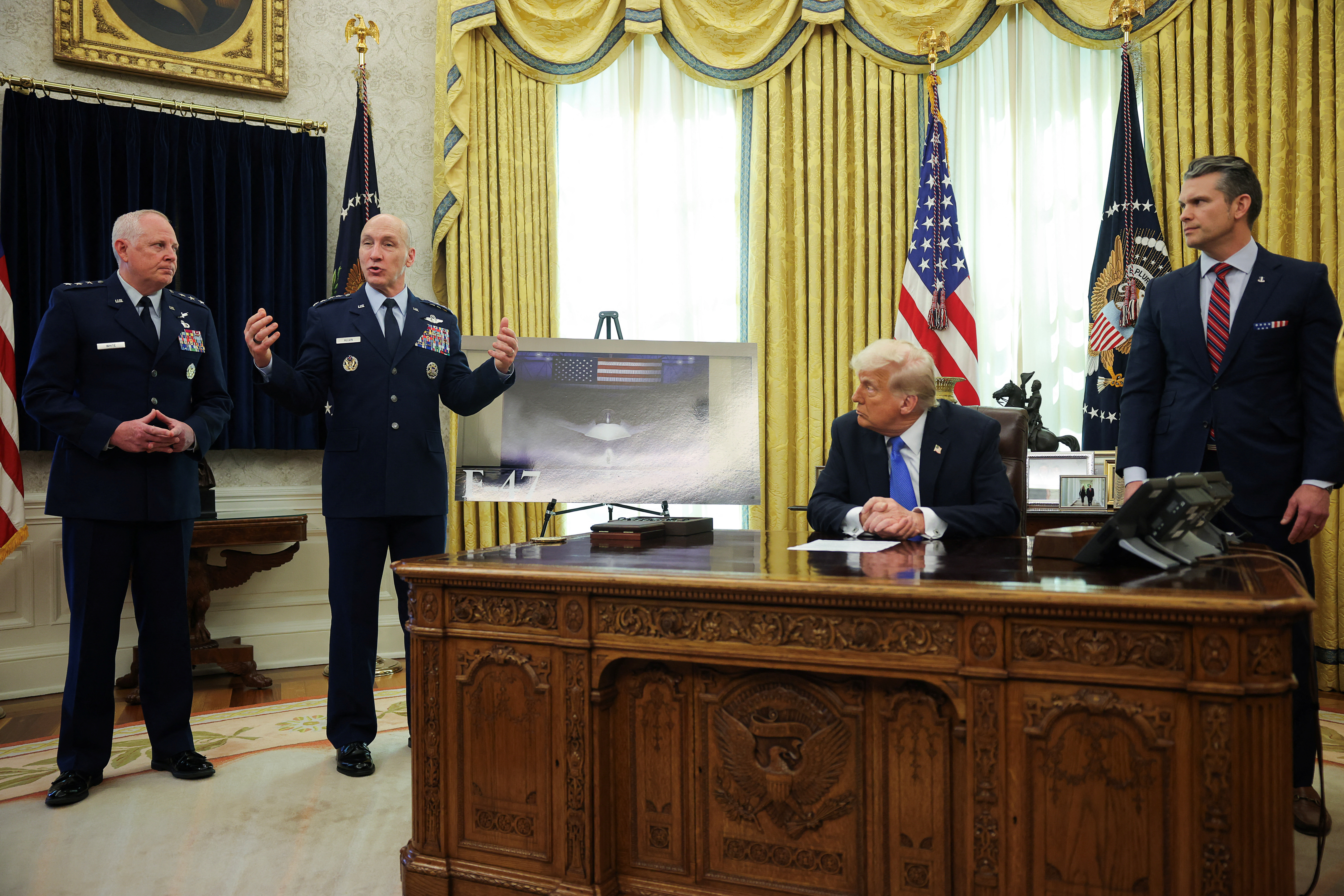 Two representatives from the Air Force give a presentation next to Donald Trump, who sits behind the Resolute Desk