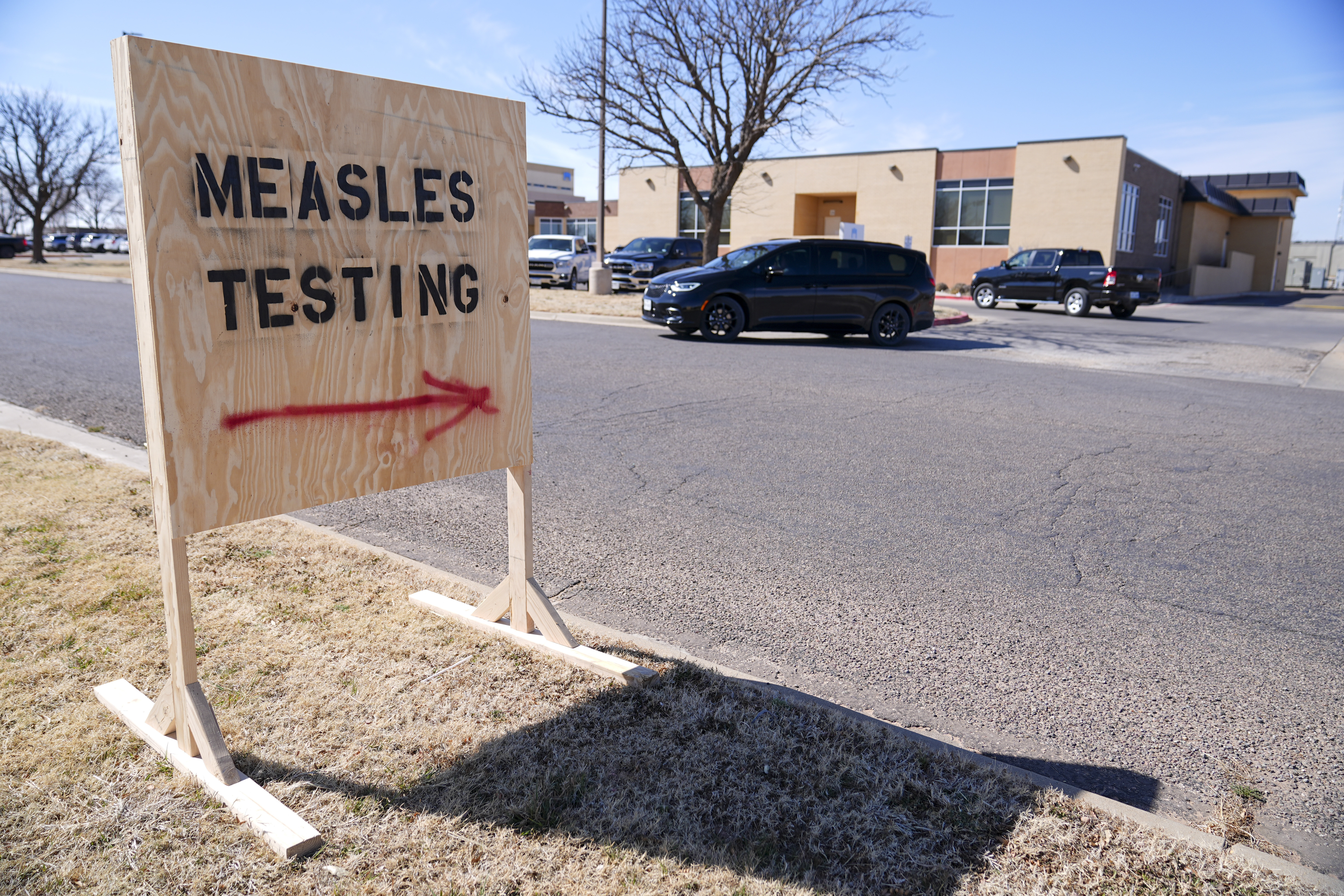 A wooden sign with a red arrow reads, "Measles testing," next to a roadway.