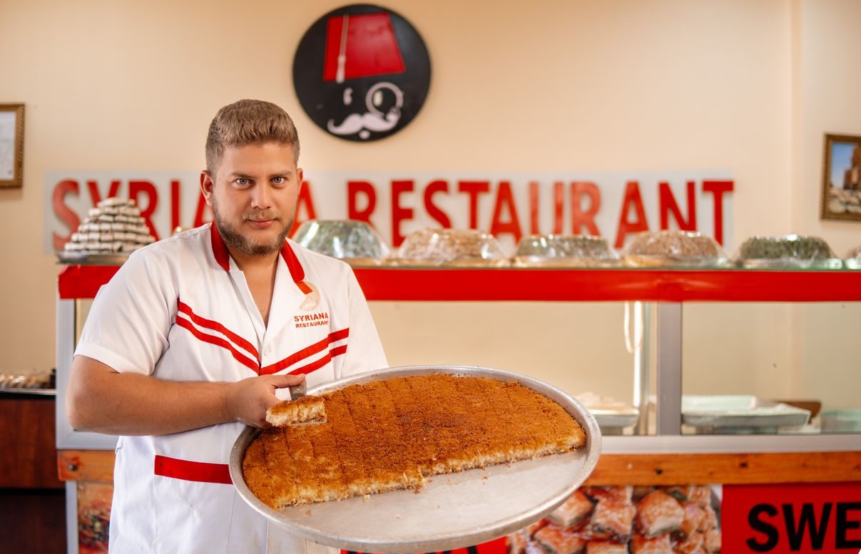 Ahmed stands holding a tray of kunafeh and smiling
