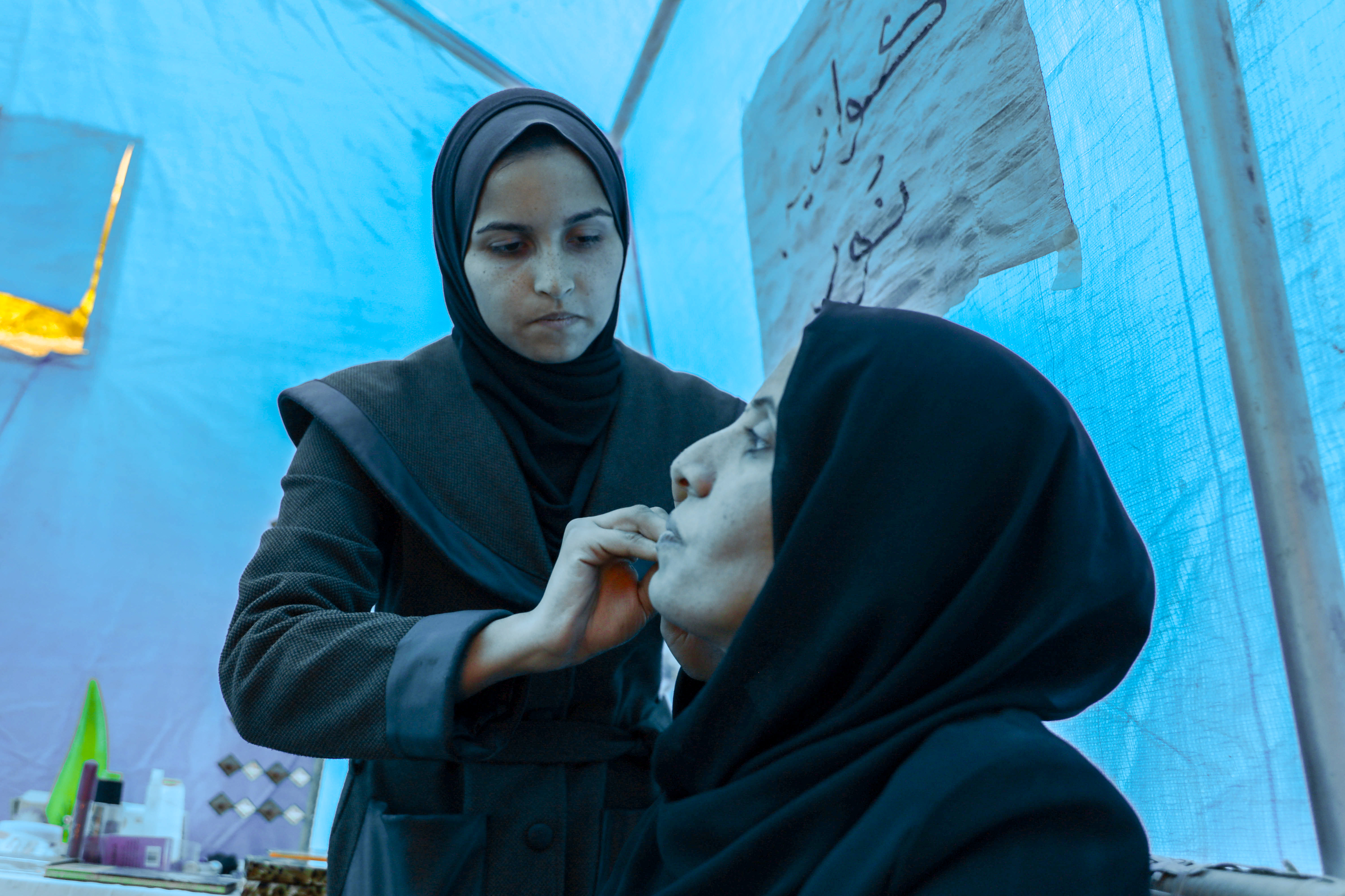 Noor works on Amani's face in the light filtering through the blue tent walls