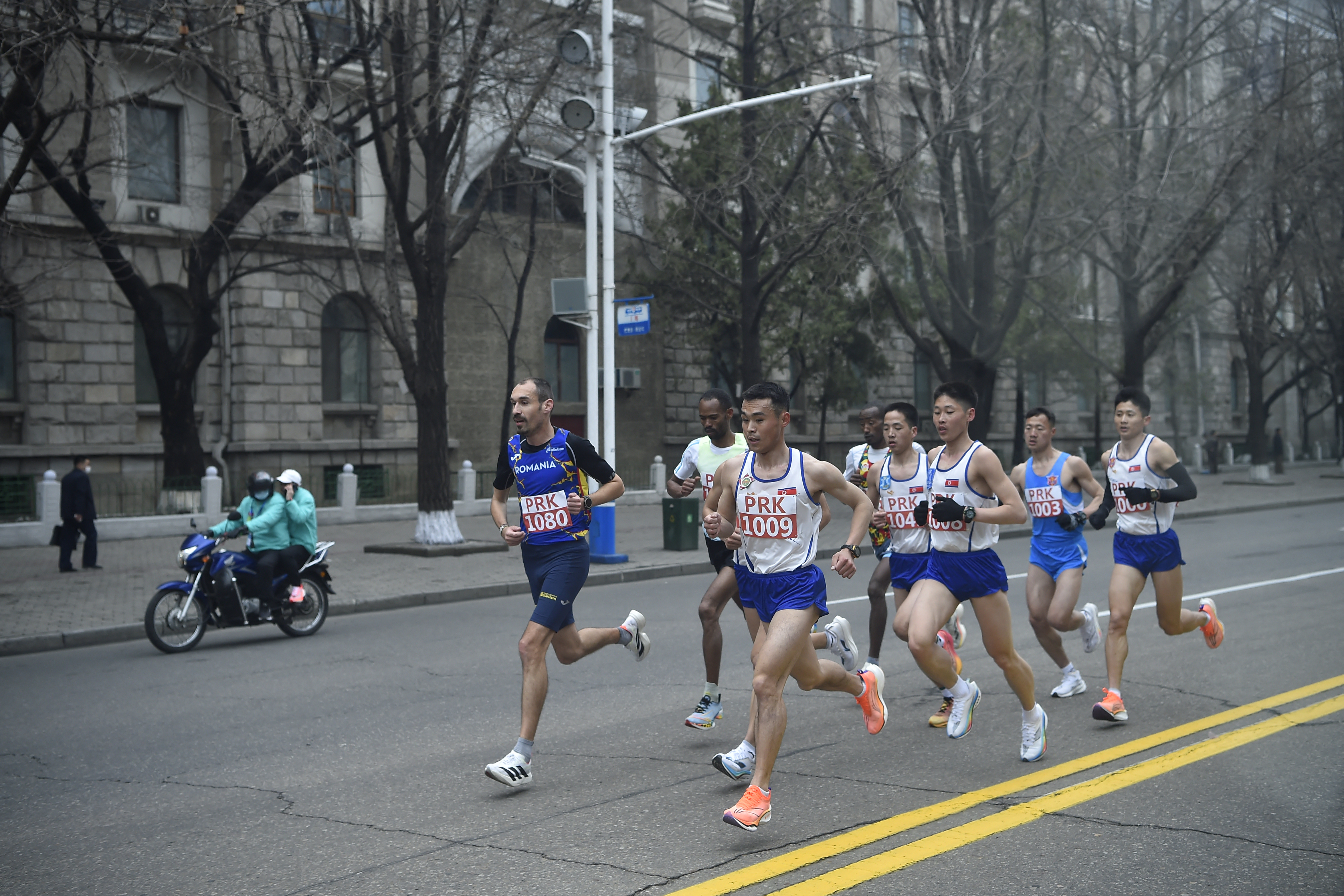 Runners wear blue and white. There is a motorcycle riding on the side of the road next to them.