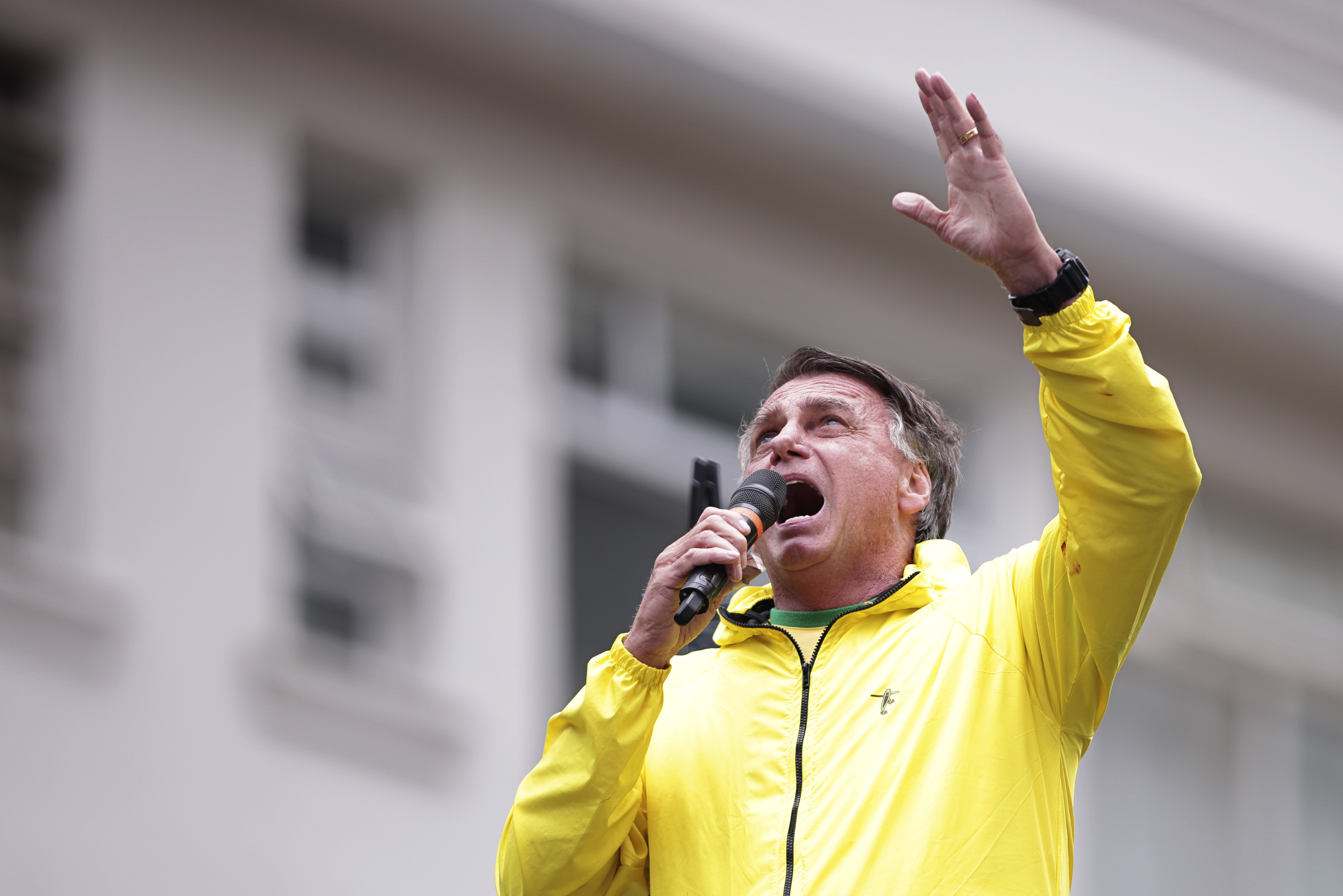 Jair Bolsonaro speaks in to a microphone and reaches upwards with his free hand