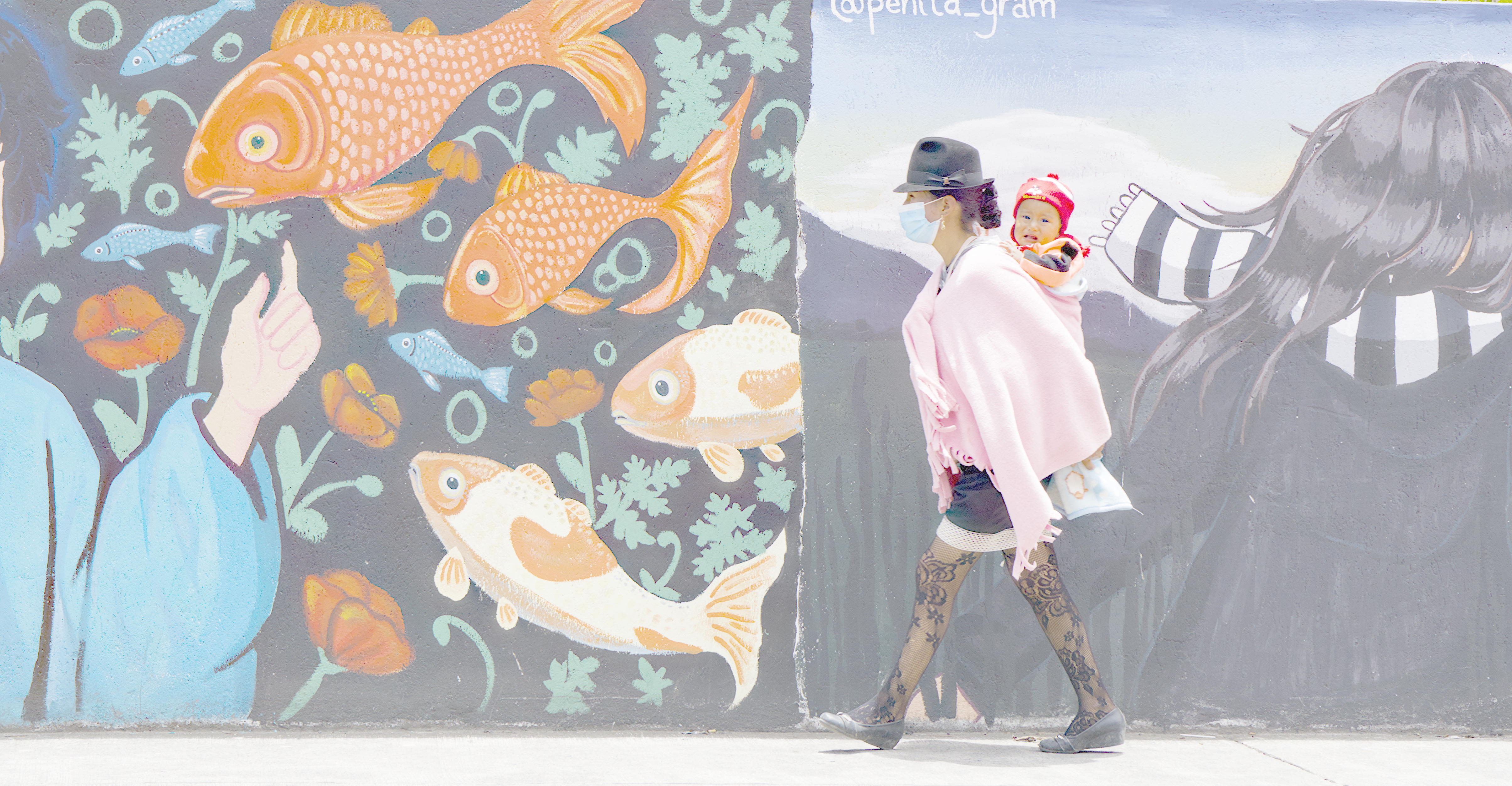 An Indigenous Ecuadorian woman walks by a mural of fish with a baby on her back.