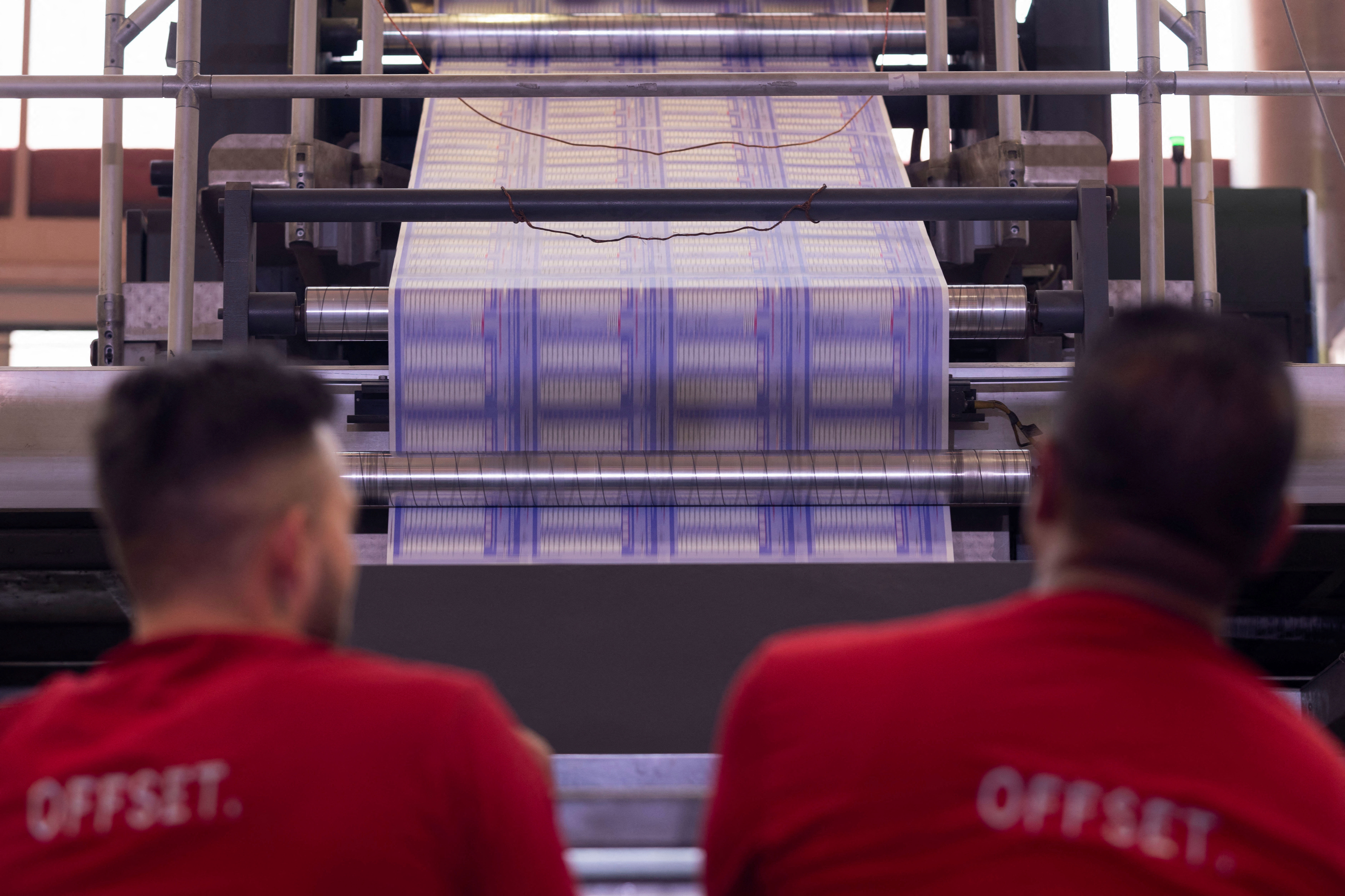 Two workers watch as machinery prints reams of ballots.