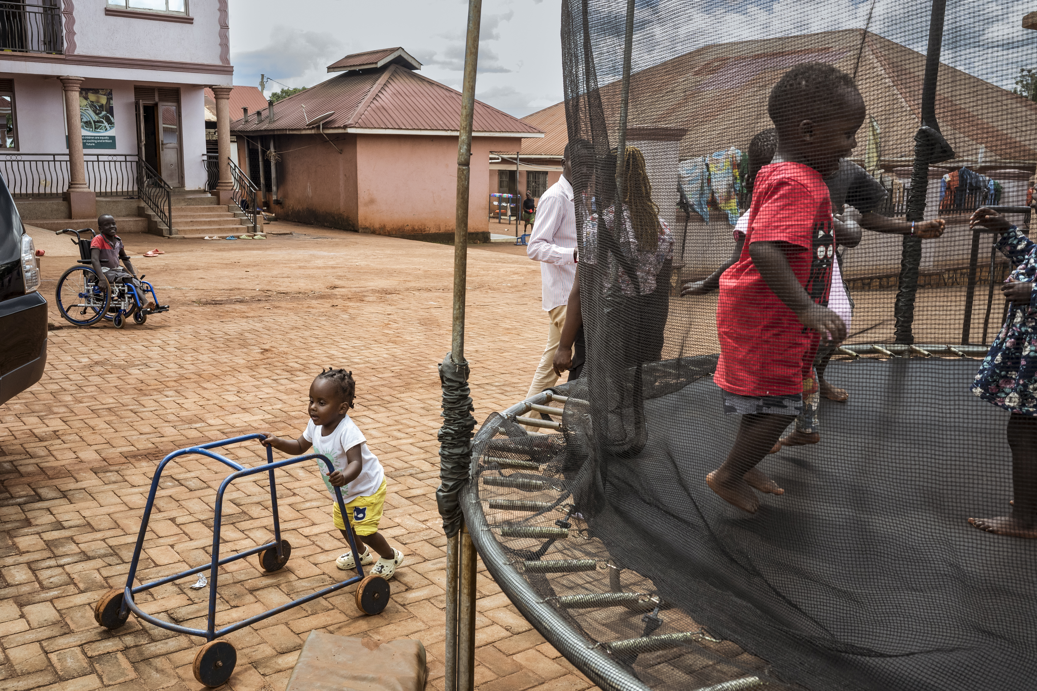 Bugembe, Jinja District, Uganda. As two-year-old Mildred, who lives with Down Syndrome, walks assisted by a walking frame, children of staff play unassisted on a trampoline at the Home of Hope. Every Tuesday is therapy day at the Home for children from the region living with disabilities. Approximately 25 parents bring their child on a first-come first-serve basis whereupon the children receive therapy from a physiotherapist and Occupational Therapist, Eddie Opio (seen here speaking with Mildred's mother Ida). The day provides the children with a required service free of charge, but has also created a community for parents of children living with disability to speak to others who are living with similar issues. [Christopher Hopkins/Al Jazeera]