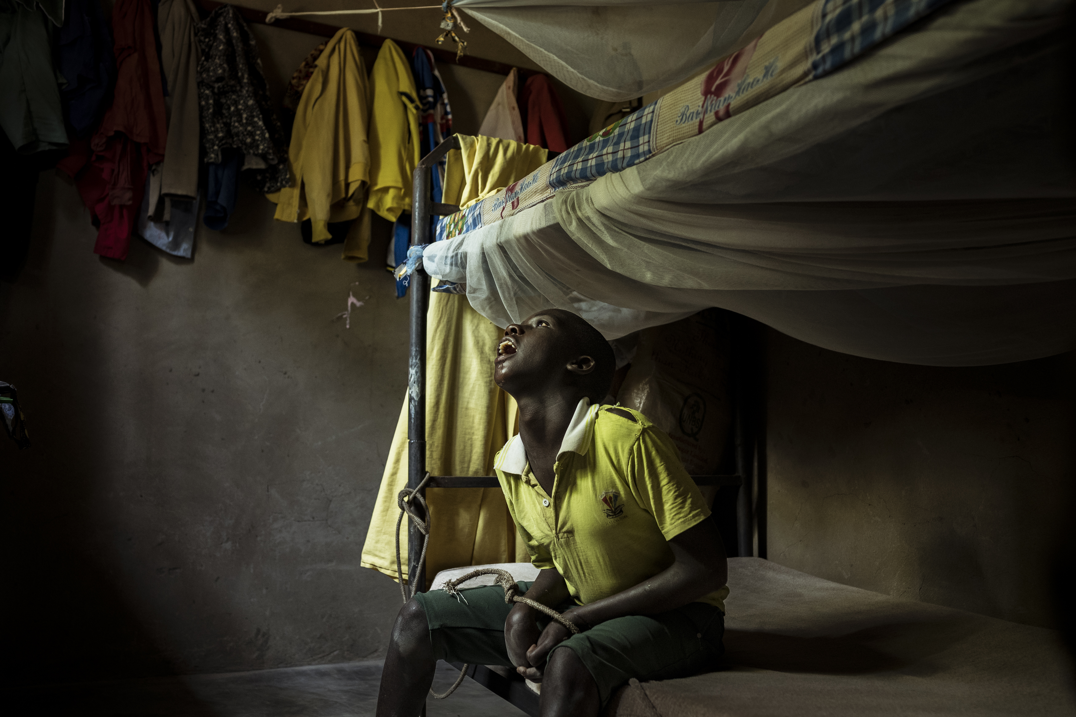 Omalera, Soroti District, Uganda. 17 -year-old David Emwodu sits hands bound by a rope and tied to the uprights of the bunk bed he shares with his brother. 