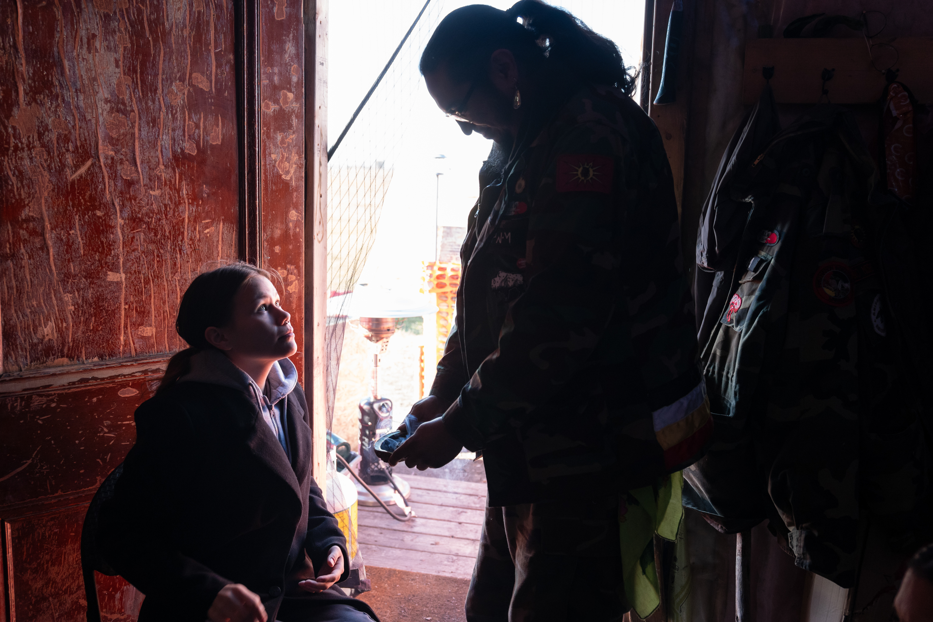 Elle Harris sits in front of a wooden door as a man stands in front of her offering to smudge her