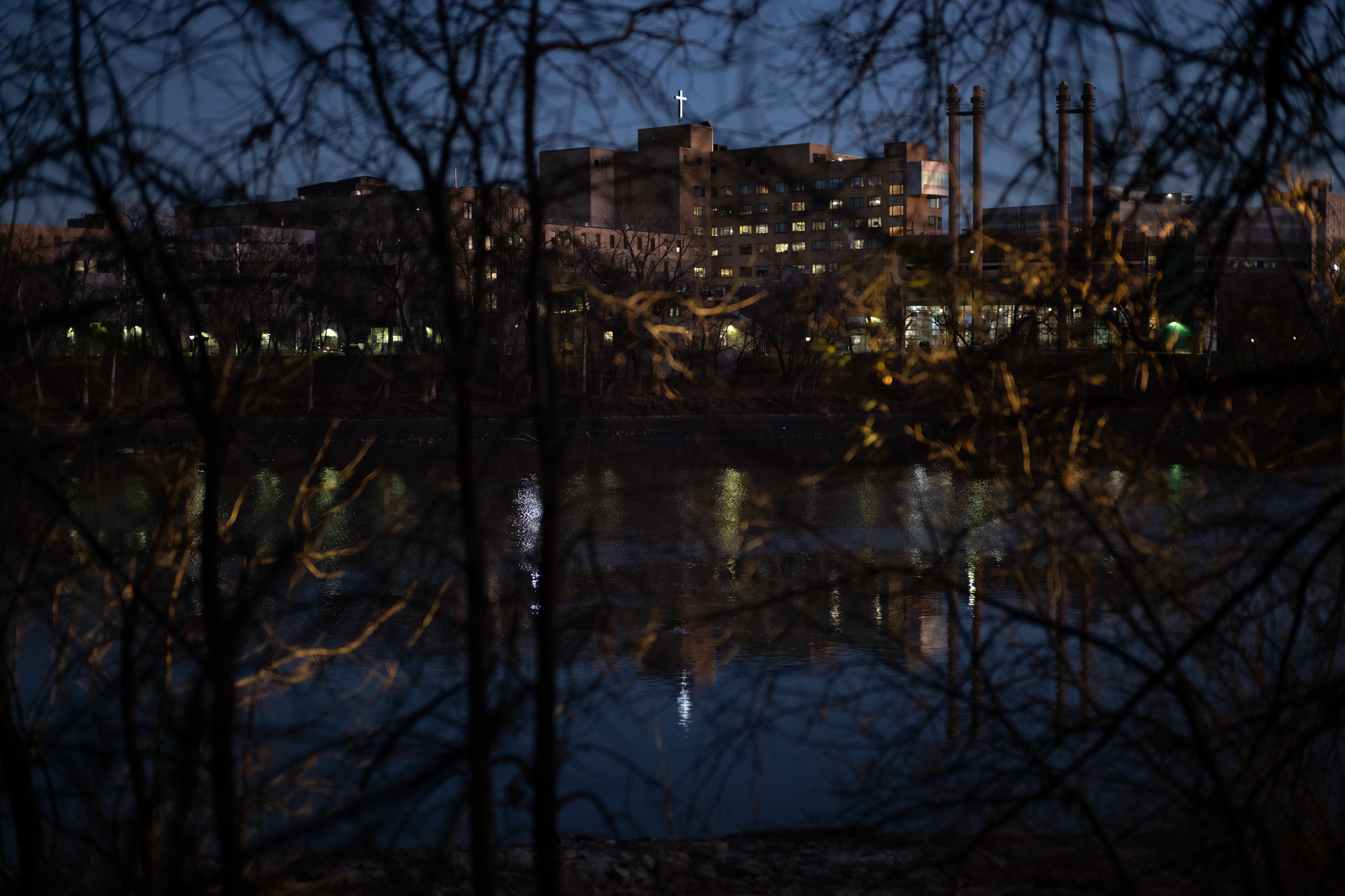 A view across the Red River at night shows tree branches in the foreground and buildings on the other side of the river