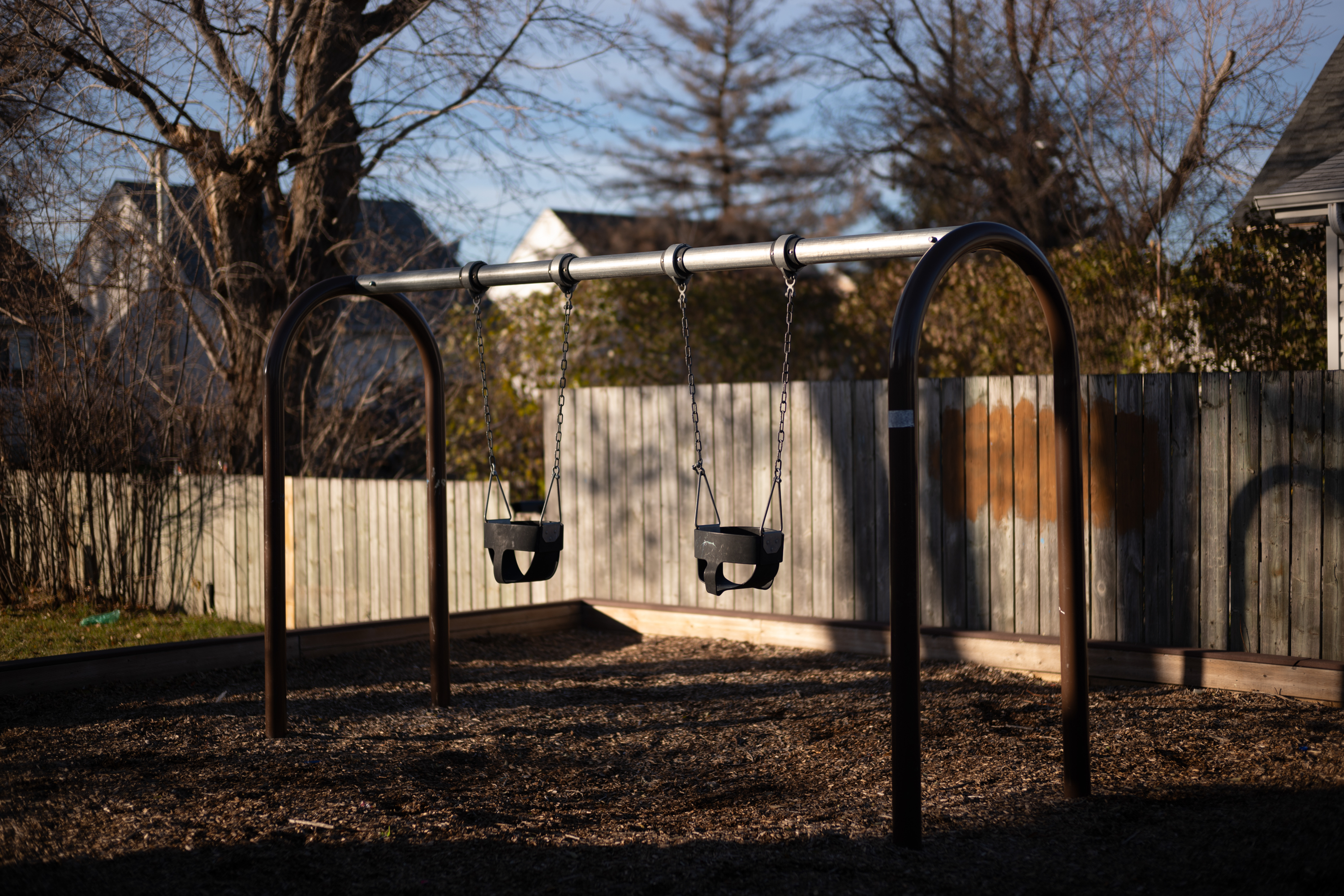 A set of swings in a park with a wooden fence behind and some trees