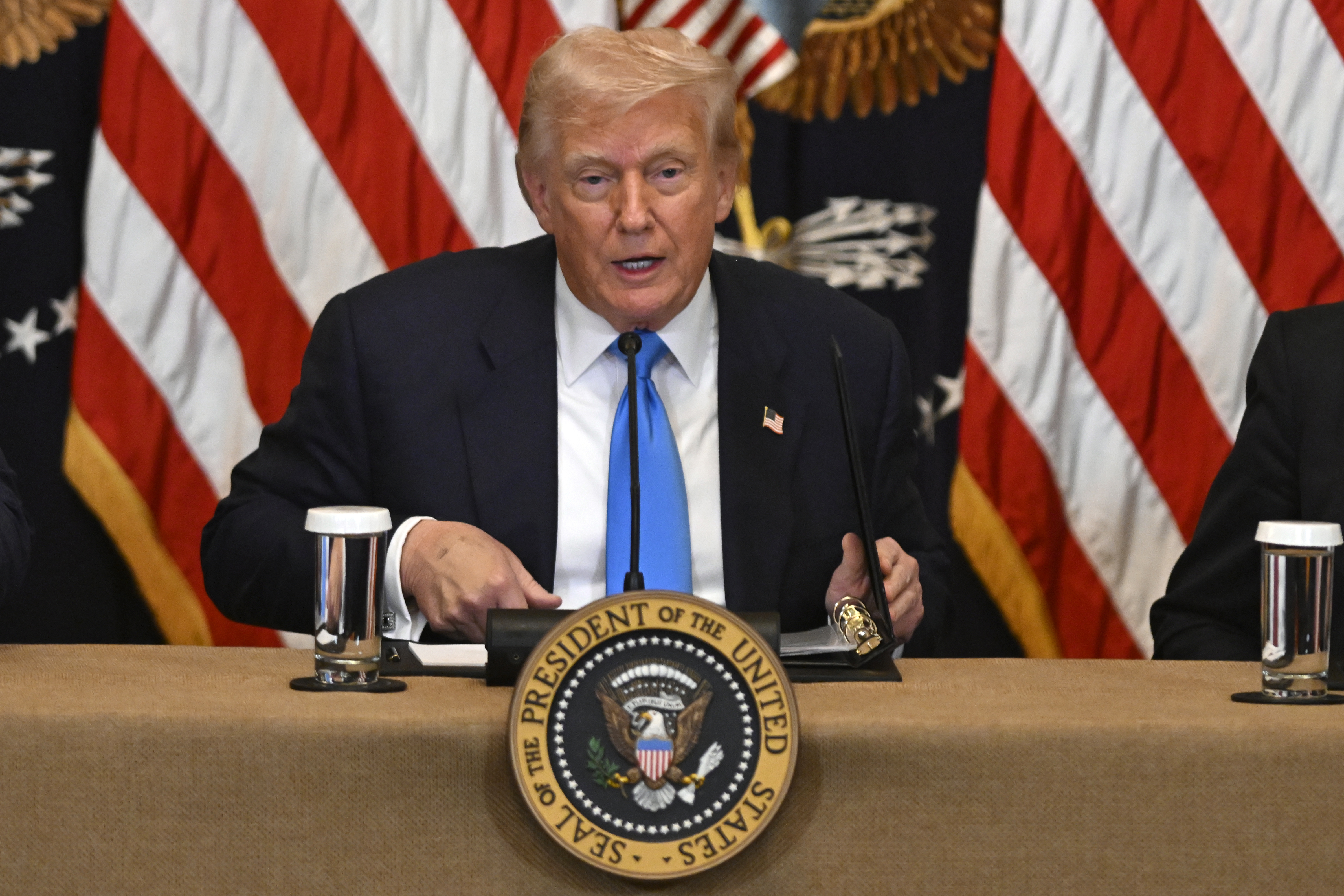 Donald Trump sits at a panel desk behind a presidential seal and in front of a row of US flags.
