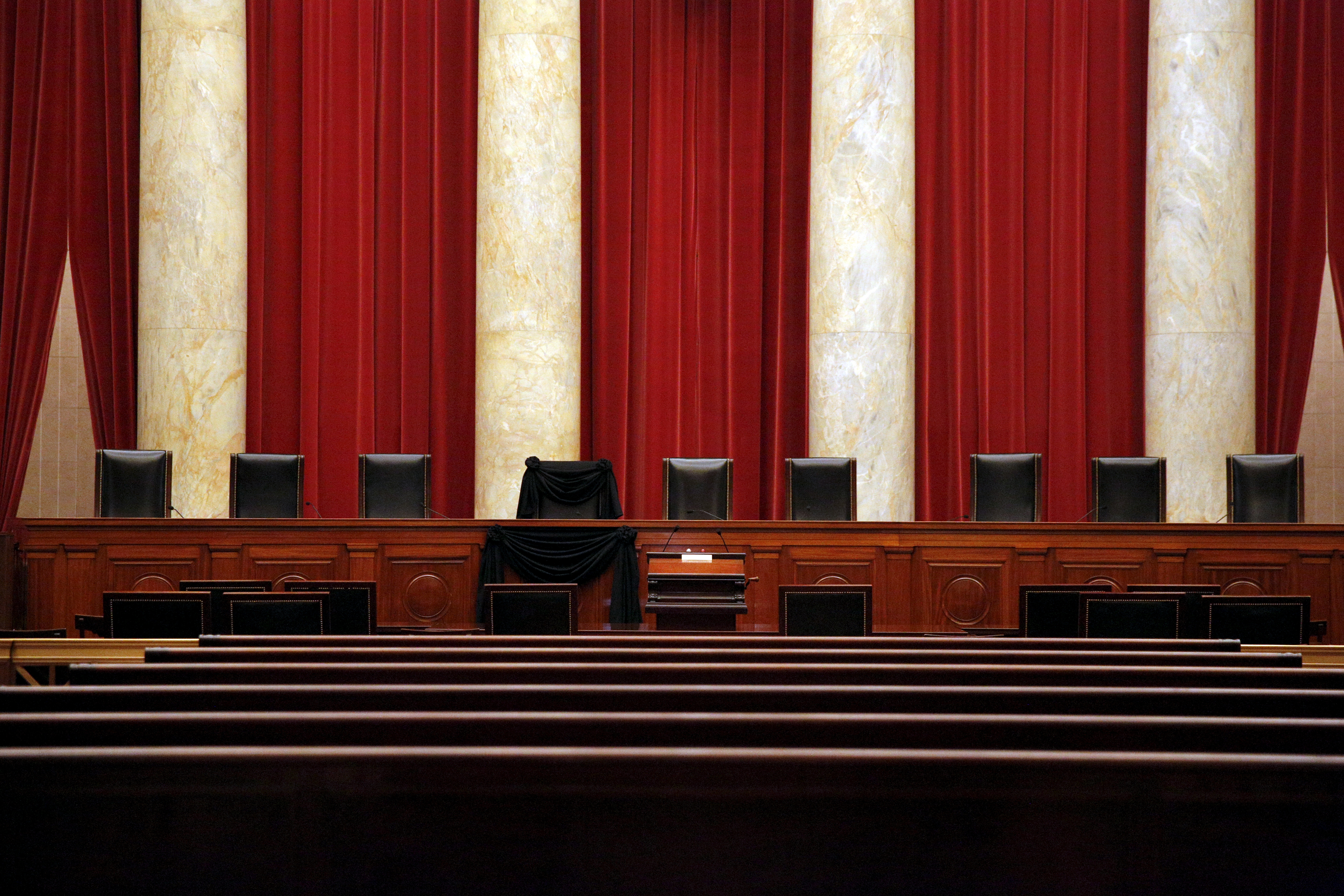 The Supreme Court's bench: a row of high-backed black seats in front of marble columns and red curtains.