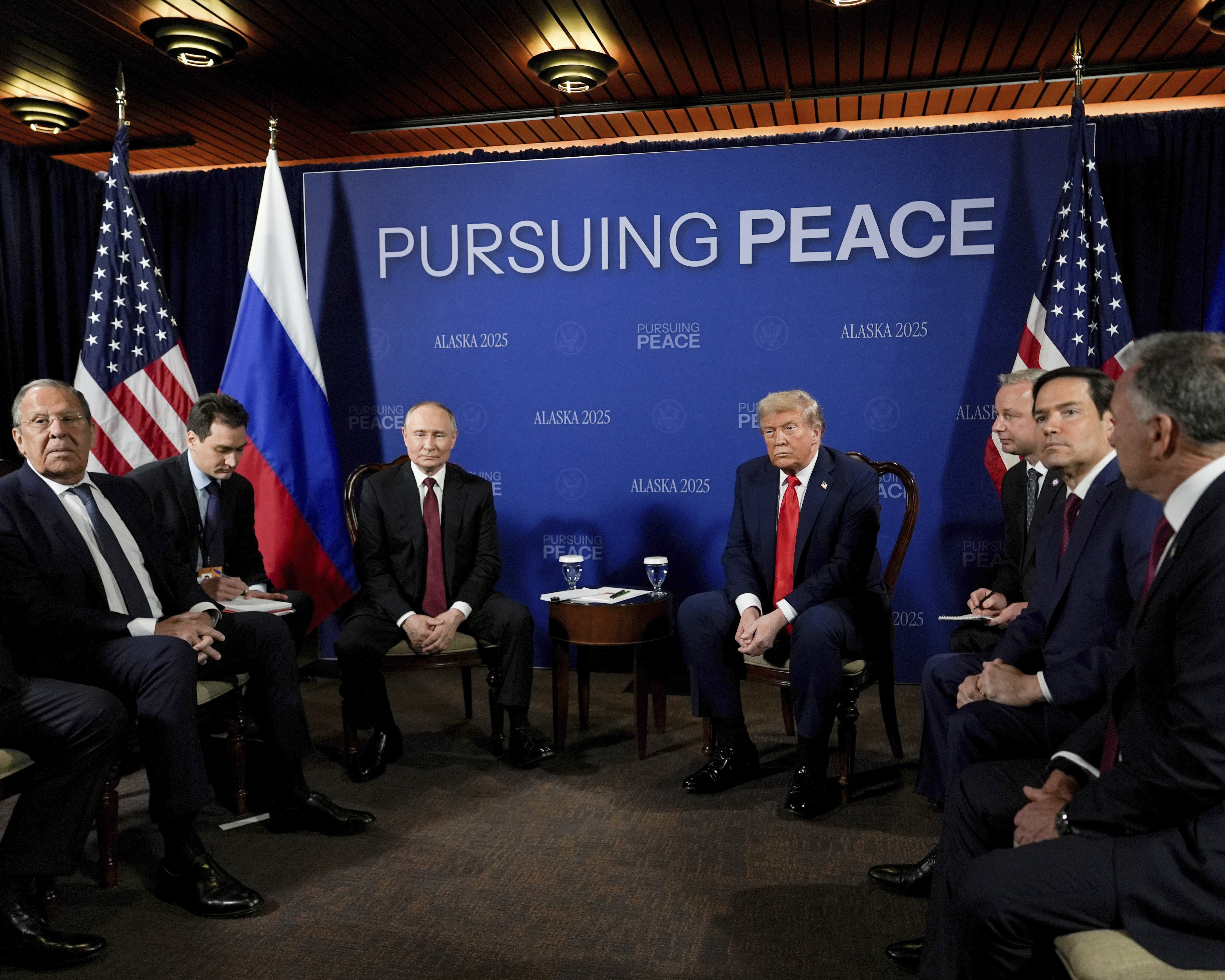Trump, Putin and members of their governments sit down together under a banner that reads "Pursuing Peace"