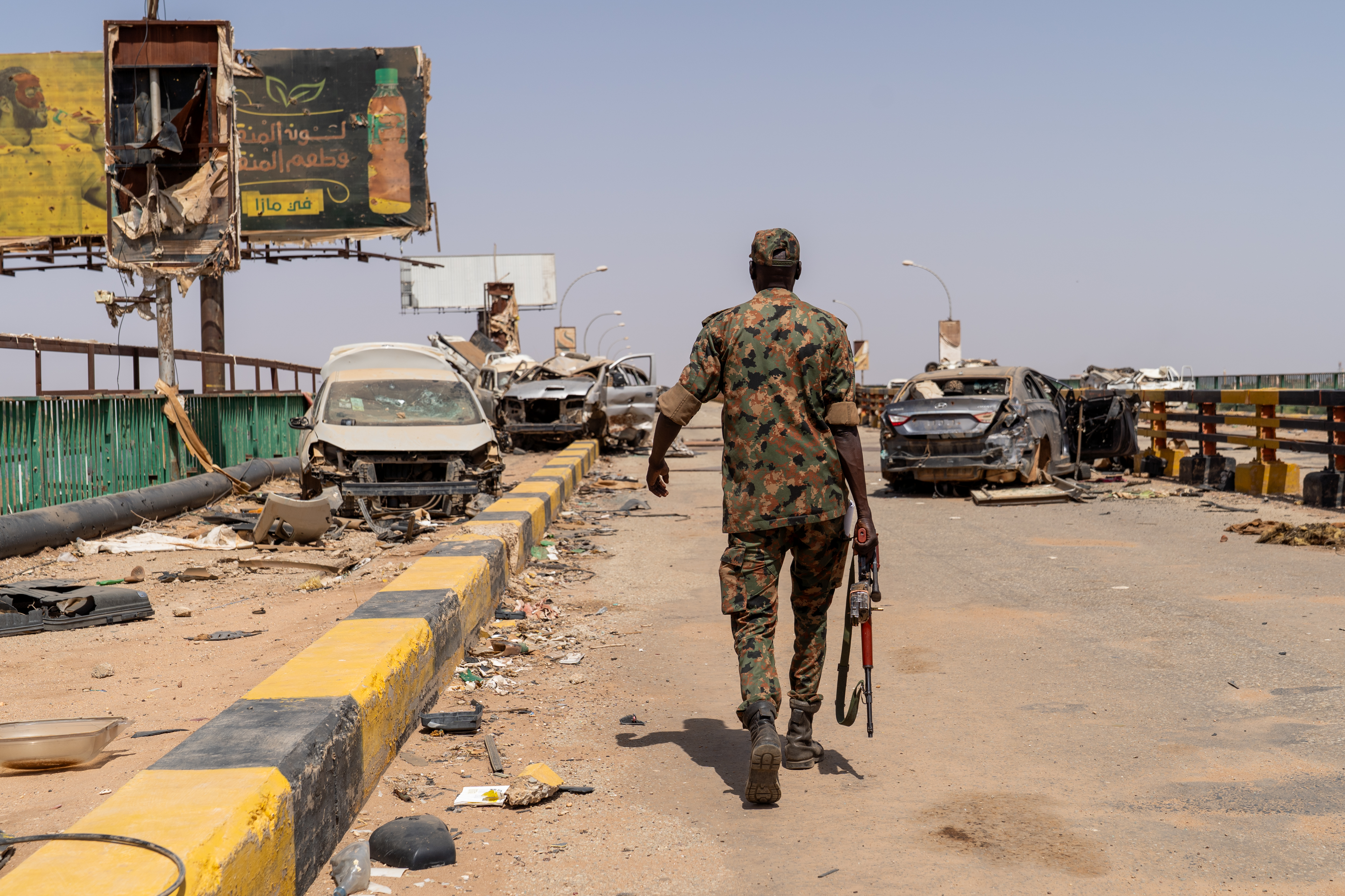 A Sudanese Armed Forces soldier walking on a bridge amidst ruins and destroyed cars.
