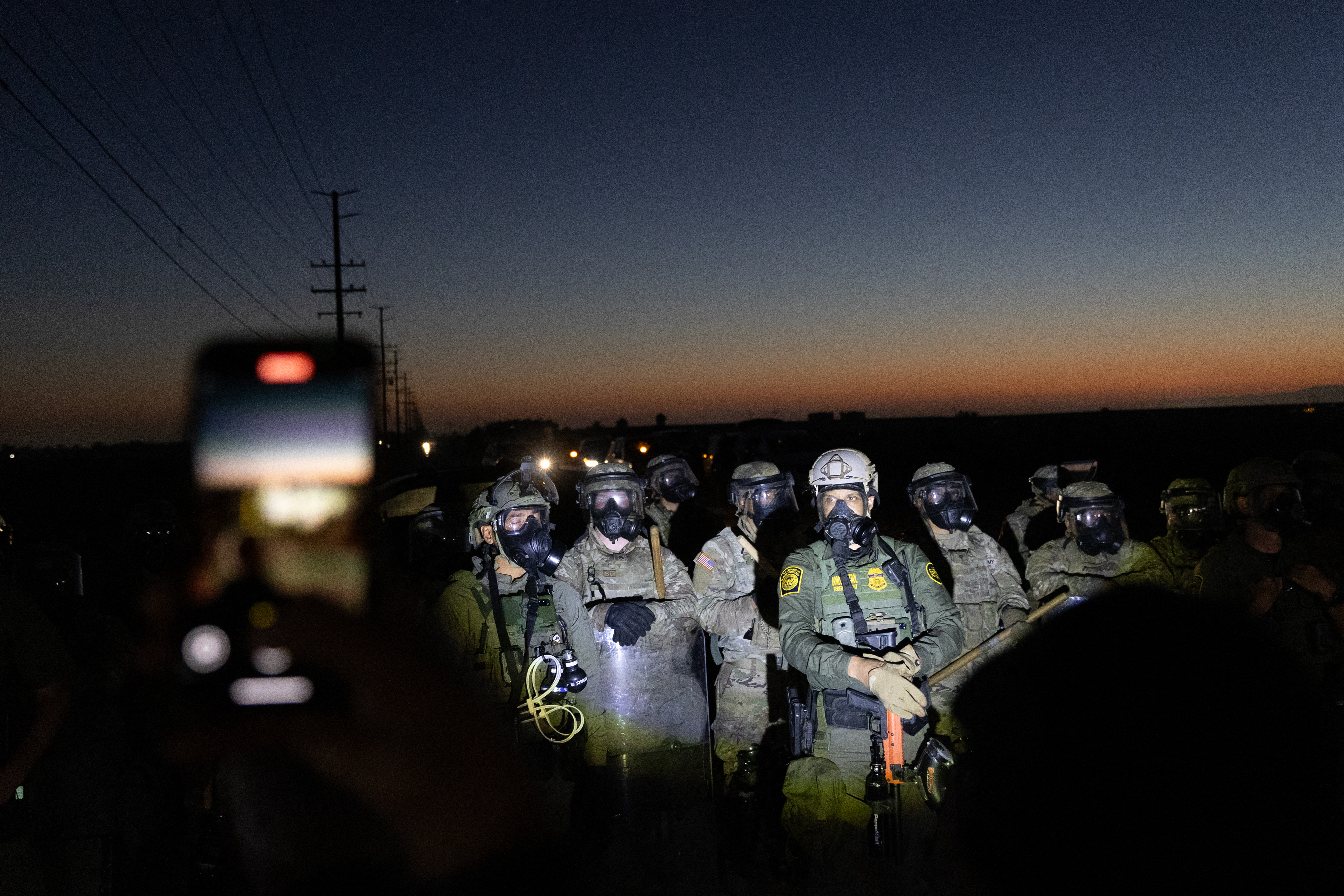 Customs and Border Protection officers and California National Guard troops hold the line as protestors shine flashlights on them after Federal Immigration agents conducted a raid on Glass House Farms in Camarillo, California, on July 10, 2025. Federal immigration agents—backed by CBP, Border Patrol units, National Guard troops, helicopters, and military style vehicles—raided Glass House Farms in Camarillo early July 10, 2025, targeting state licensed cannabis and other crop operations, detaining between a dozen and 100 farmworkers, and deploying smoke or tear gas canisters and rubber bullets to disperse a crowd of several hundred protesters on Laguna Road. (Photo by BLAKE FAGAN / AFP)