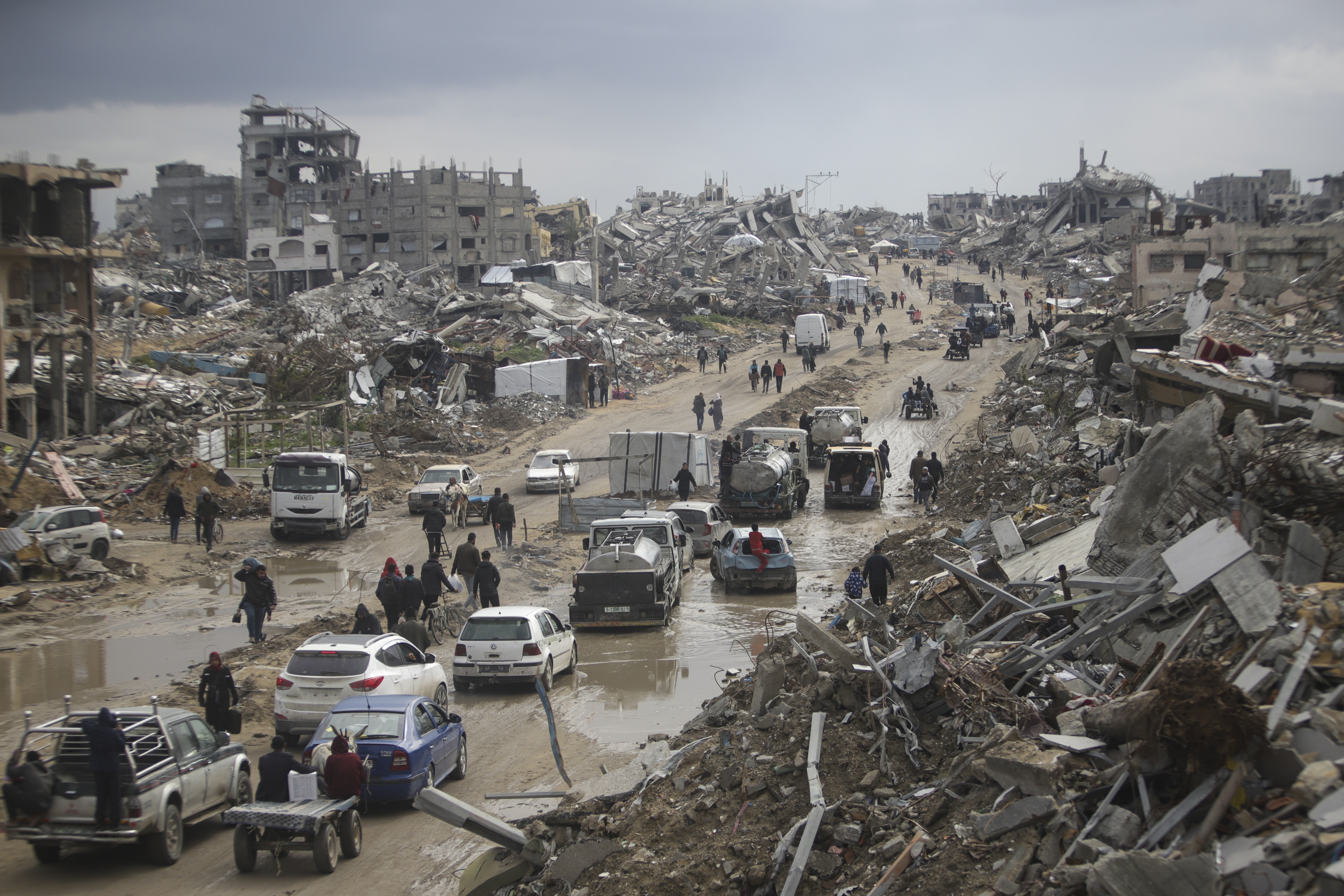 Cars and pedestrians move along a road amid widespread destruction.