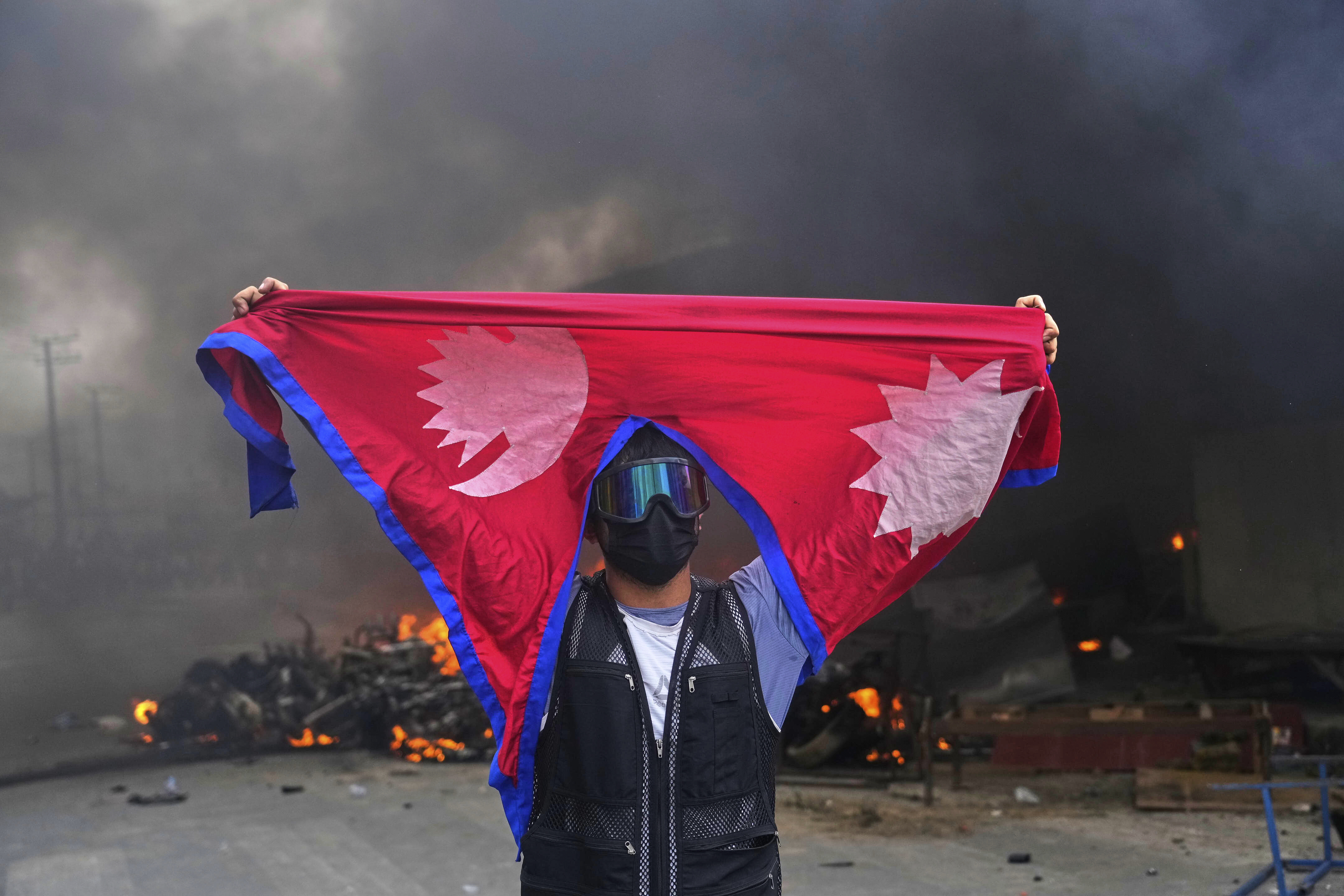 A protester shouts slogans, carrying the national flag, with a fire burning in the background.