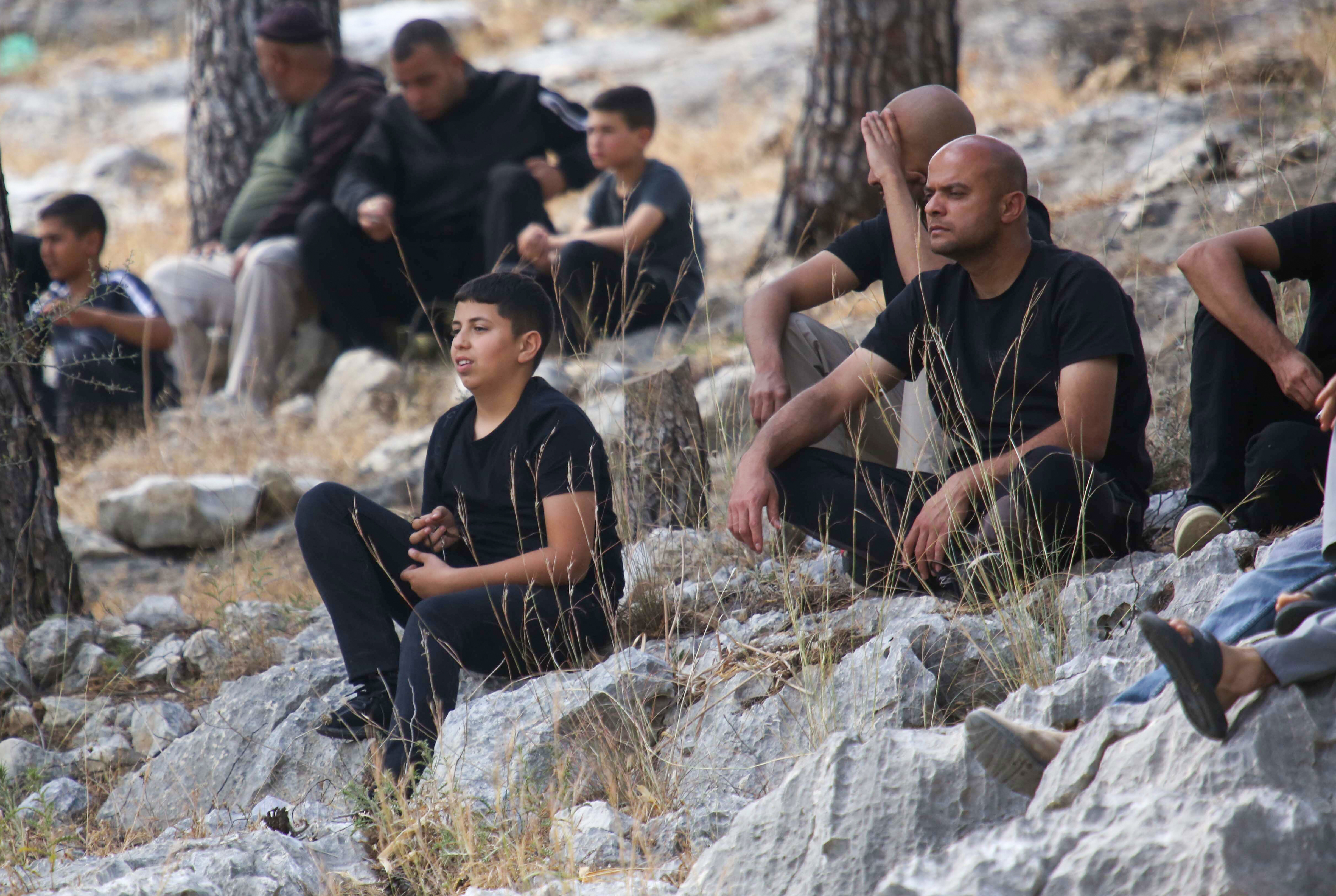 Men and young men sitting on rocks looking aghast off to one side at the ongoing demolition