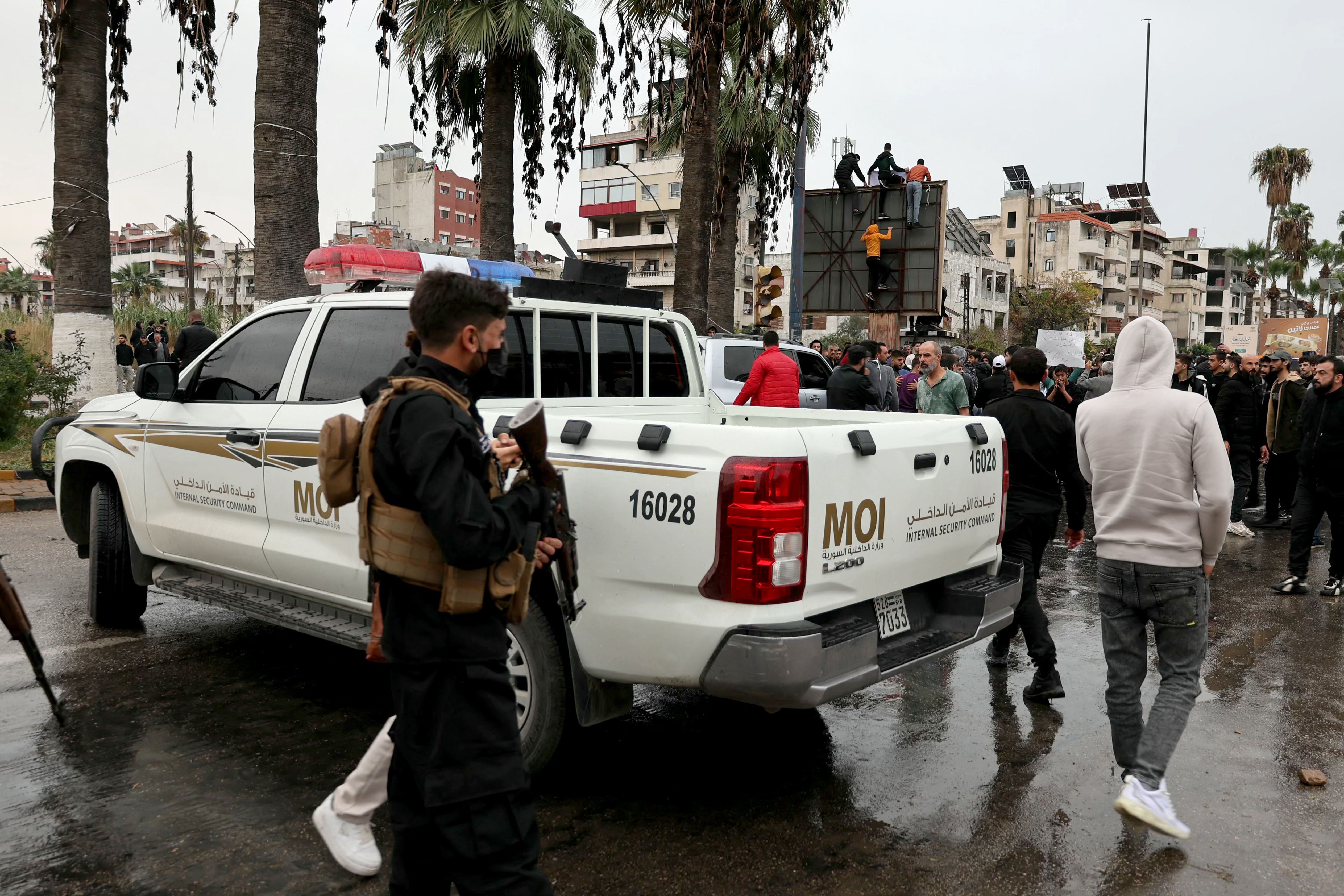 Syrian Internal Security officers stand near their vehicle, as people take part in a protest in al-Azhari Square, following recent attacks against the Alawite minority community, an offshoot of Shia Islam, in the coastal city of Latakia, in Syria's Alawite heartland on November 25, 2025.