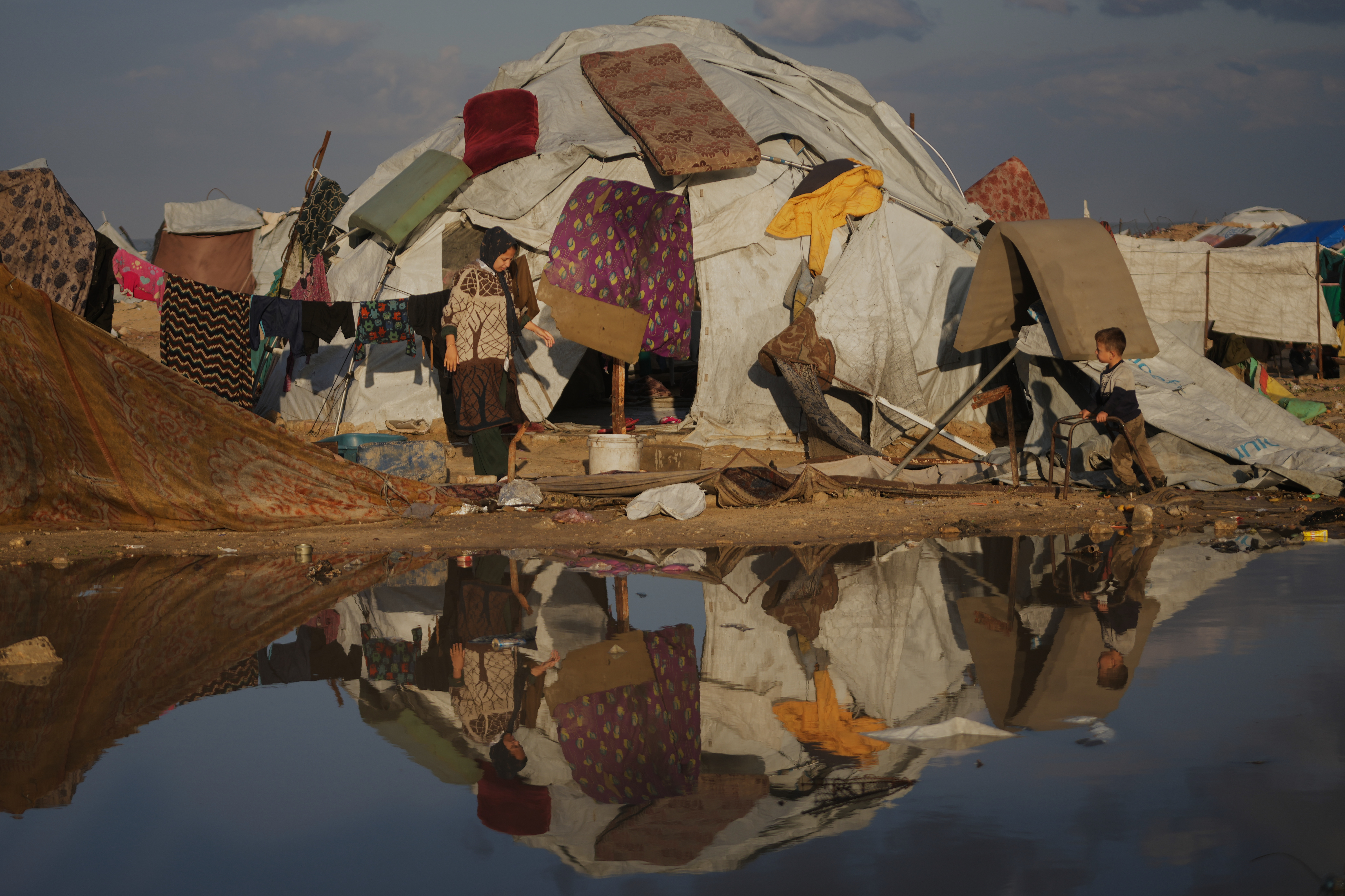 A woman and her nephew stand in front of their family's tent as mattresses and clothes hang to dry in a makeshift camp for displaced Palestinians set up on the beach in Gaza City, Tuesday, December 16, 2025. [Abdel Kareem Hana/AP]