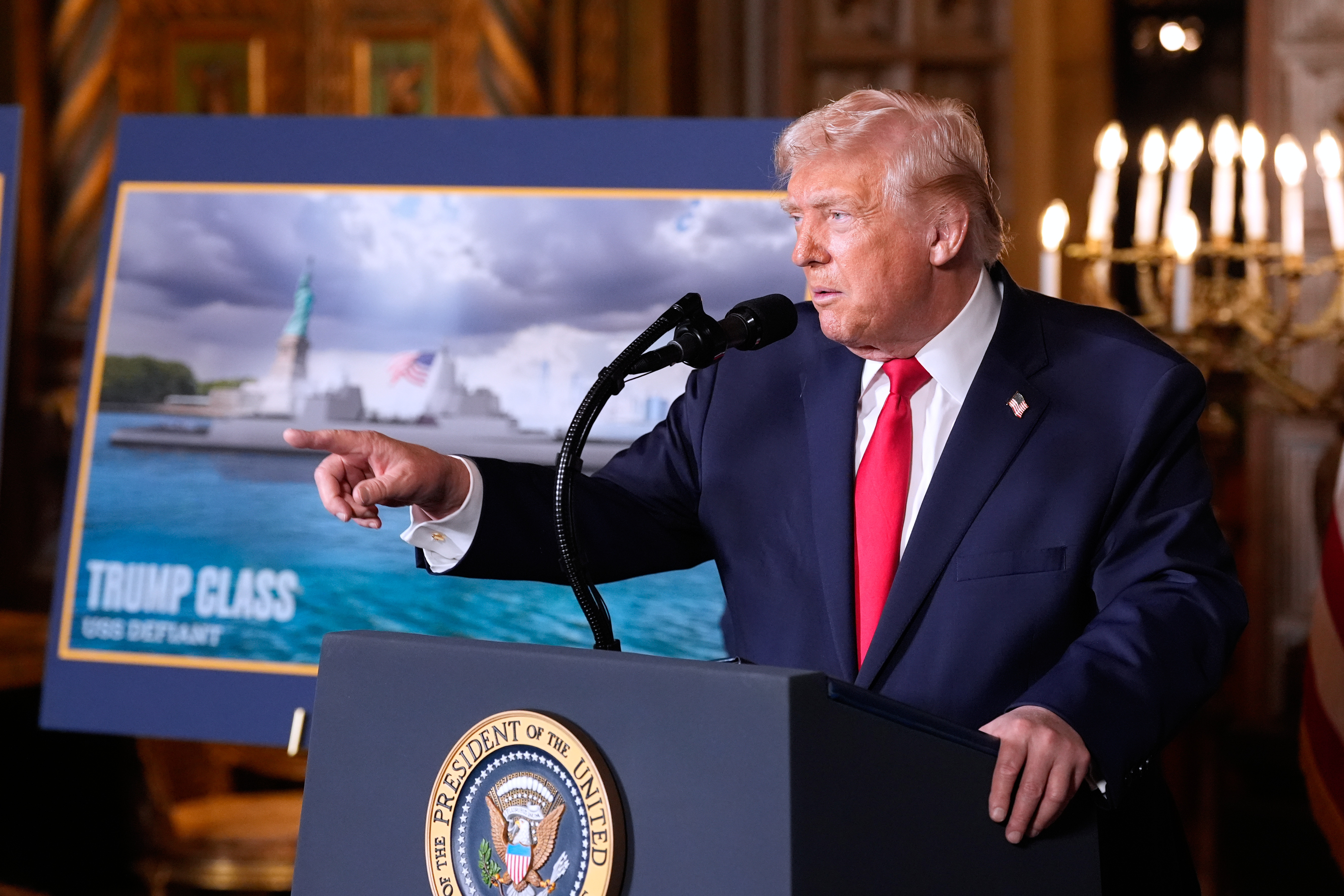 Trump speaks at a podium in front of a sign for "Trump Class" ships
