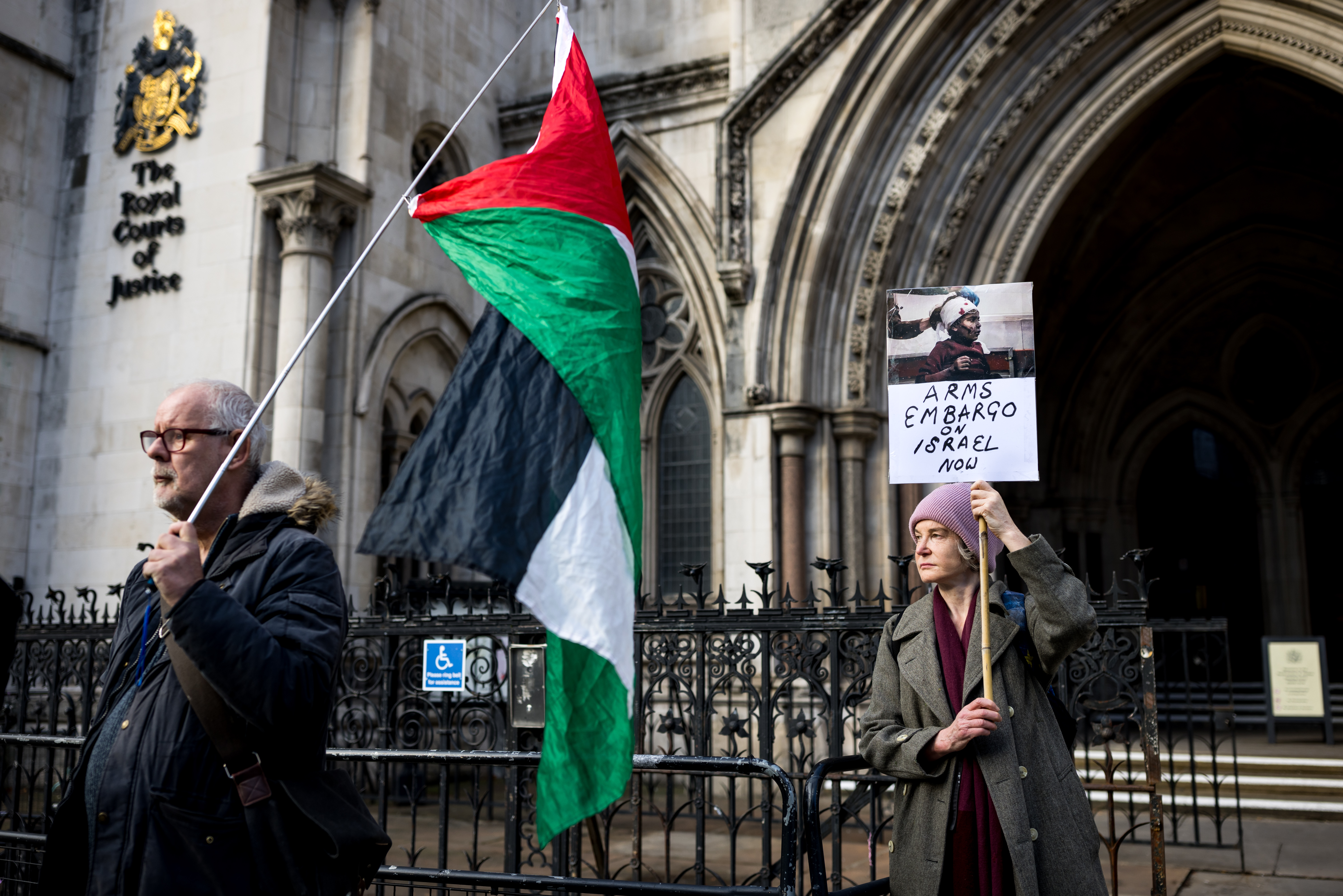 Protesters holding a placard and a Palestinian flag gather outside a court.