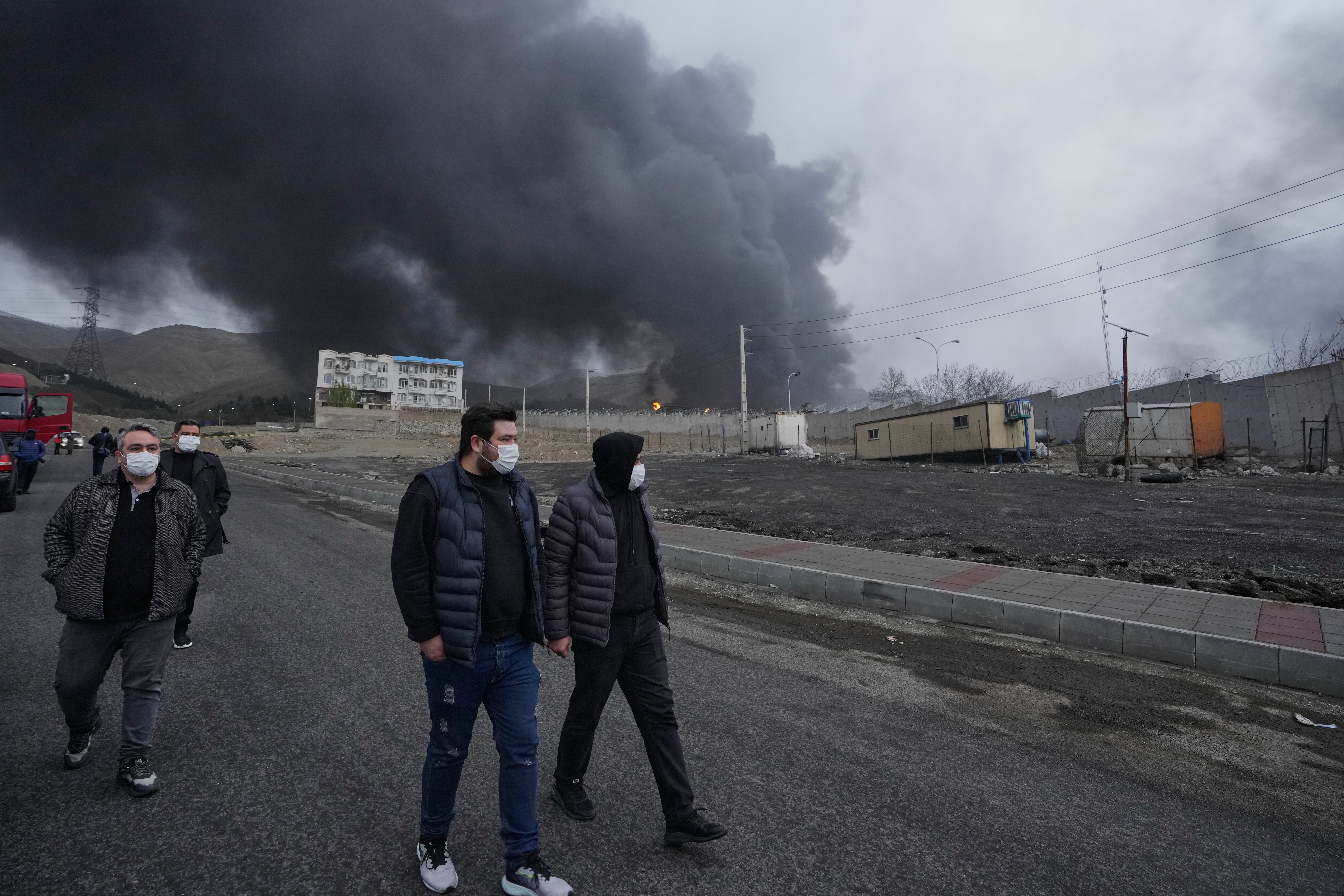 Four men walk as a thick plume of smoke from a U.S.-Israeli strike on an oil storage facility late Saturday lingers in the cloudy sky behind them in Tehran, Iran, Sunday, March 8, 2026. (AP Photo/Vahid Salemi)